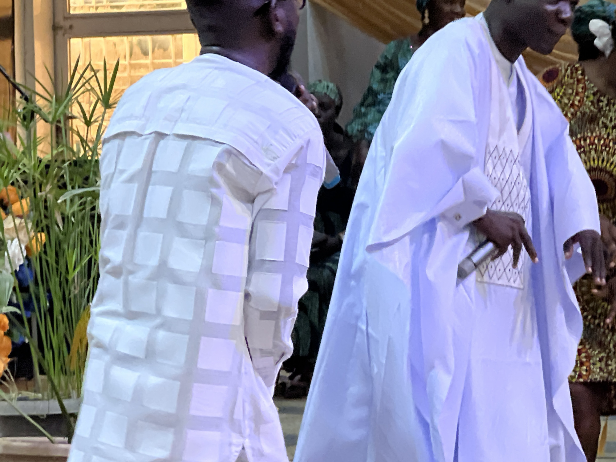 The image captures a close-up, interior scene featuring individuals in traditional attire. In the foreground, a person is seen from the back, wearing a white garment with a checkered pattern of light-colored squares. To the right, another individual is partially visible, adorned in a flowing light blue robe, holding a microphone. The background reveals a window with light filtering through, and behind the people, there are hints of other individuals in patterned clothing and decorative elements. The presence of plants on the left suggests a decorative arrangement within the space. The overall ambiance appears to be that of a gathering or event, likely within a community hall or similar venue in Kabong, Nigeria.
