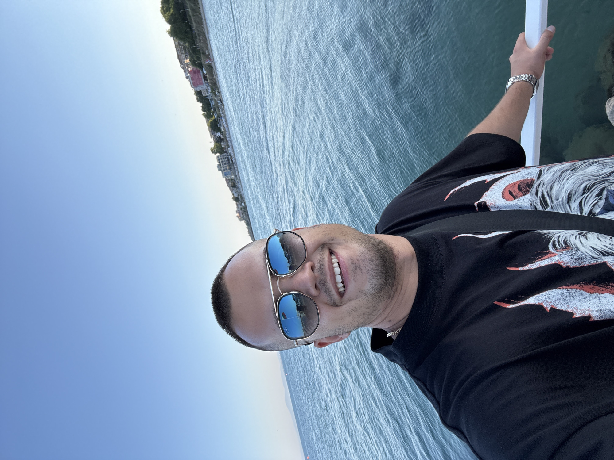A man is taking a selfie outdoors, with a body of water and a coastline in the background. The man is wearing a black t-shirt with a graphic print, sunglasses, and a watch. He has short dark hair and a beard, and he is smiling. The sky is clear and blue, suggesting it is daytime. The water is calm and appears to be the ocean. The coastline shows some buildings and greenery. The man's arm is visible, resting on a white railing, and he is wearing a silver watch. The overall mood is casual and relaxed.
