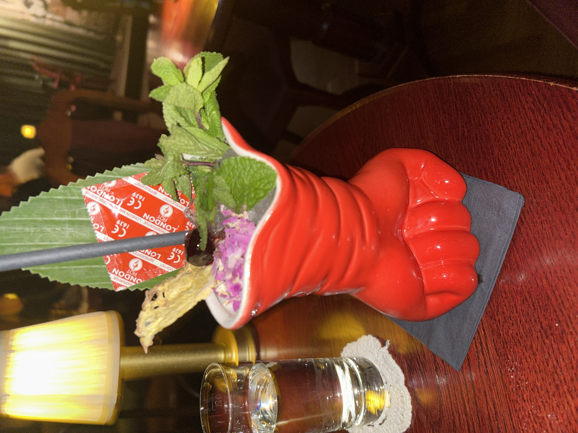 A cocktail is served in a red, fist-shaped ceramic glass, garnished with mint, a dried flower, and a leaf. The glass is placed on a dark gray napkin on a dark wooden surface. The cocktail also includes a red packet of "London" brand condoms attached to a straw. A small glass of liquid sits on a small white crocheted coaster next to the cocktail glass. The scene appears to be set indoors, possibly at a bar or restaurant, with a partial view of furniture and lighting elements in the background. There is no indication of the location being in Berlin, Germany.