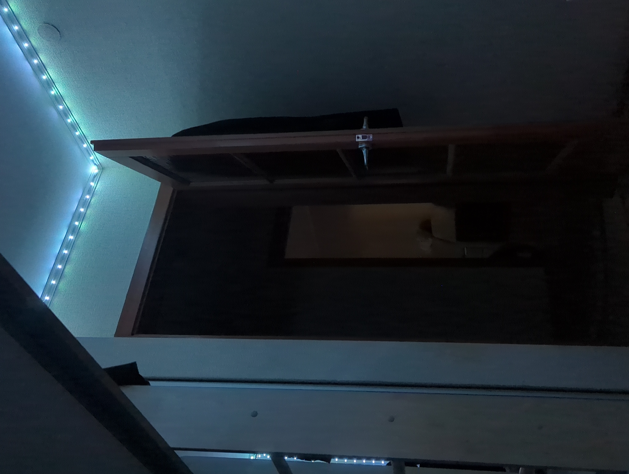 The image depicts an interior scene. A wooden door is ajar, revealing a dark hallway beyond. Above the doorway, a strip of LED lights illuminates the ceiling and the wall with a blueish-green glow. A dark garment, possibly a coat, hangs on the wall near the door. To the left of the door, a section of a light-colored wall or cabinet is visible. The environment appears to be a room in a residential building, possibly a hallway or entrance area. There are no people visible in the image, and no specific identifiable landmarks to place it in Votkinsk, Russia.
