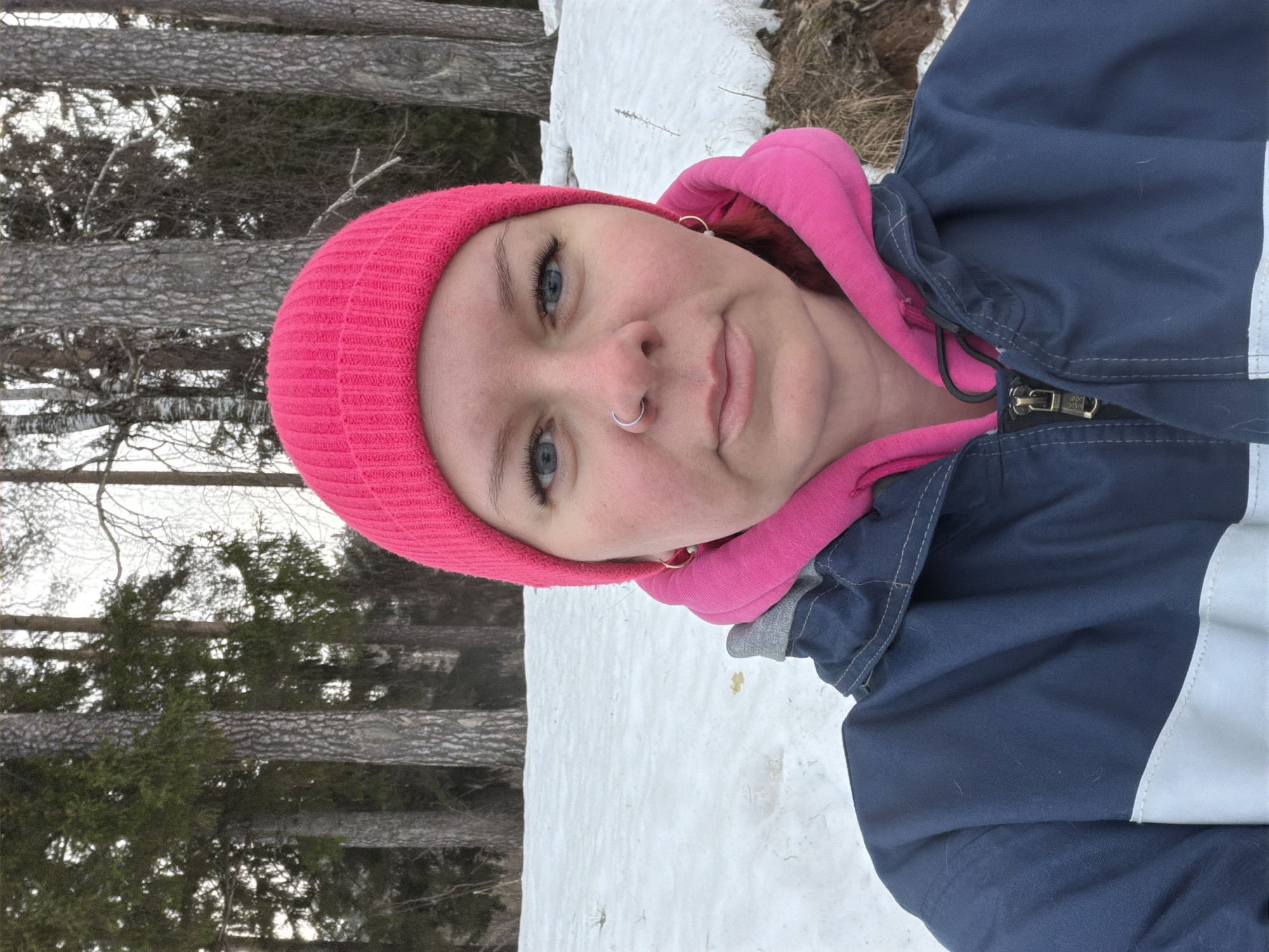 An adult female is depicted in an outdoor, chest-up portrait, looking directly at the camera. She wears a bright pink knitted beanie, a bright pink hooded sweatshirt, and a dark blue and light gray jacket with a zippered front. Her features include blue eyes with visible eyelashes, a silver hoop nose ring in her left nostril, and small gold hoop earrings. The background shows snow-covered ground with areas of exposed brown earth, indicating a winter or early spring setting. A wooded area behind her contains both coniferous evergreen trees and bare deciduous trees, including several birches. The scene is located in the city of Chaikovsky, Russia.