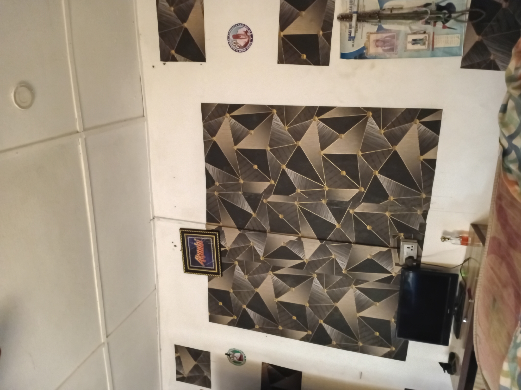 The image depicts an indoor scene, likely a room in a residential building in Ipota, Nigeria. The walls are painted white and feature a geometric patterned wallpaper in shades of grey, black, and gold. Several framed pictures and circular stickers adorn the walls. A framed picture with the word "Asante" is prominently displayed. A small television set is placed on a wooden desk below the wallpaper, next to a power outlet. Other objects on the desk include a bottle of liquid, what appears to be sunglasses, and a small black object. Part of a bed with a patterned comforter is visible in the foreground. The ceiling is white and appears to be made of suspended panels. The overall impression is that of a simple, decorated living space.