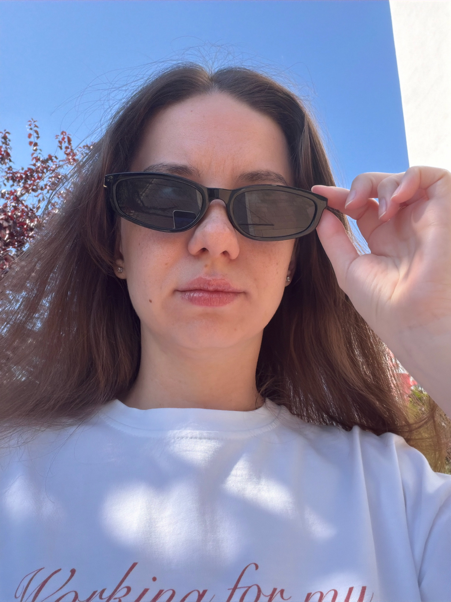 A young woman is taking a selfie outdoors under a clear blue sky. She is wearing black sunglasses and a white t-shirt with red text on it. She is holding the edge of her sunglasses with her right hand. In the background, there is a tree with red leaves and a white wall. The image appears to be taken on a sunny day. There are no identifiable landmarks to suggest a specific location within Kyiv, Ukraine.