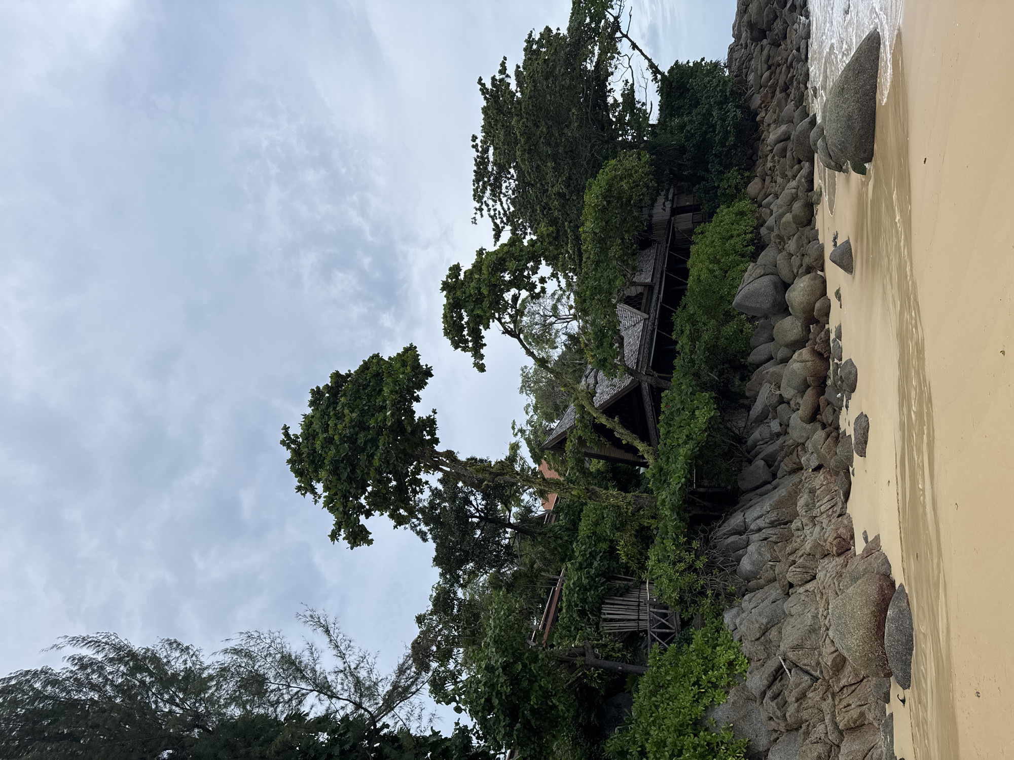 The image depicts a tropical beach scene in Karon, Thailand. The foreground is a sandy beach with gentle waves lapping at the shore. The midground is dominated by a rocky coastline covered in lush green vegetation. Interspersed within the foliage are parts of a building with a thatched roof, suggesting a resort or traditional dwelling. The background features tall trees and a cloudy sky. There are no people visible in the image. The scene is one of natural beauty and tranquility.