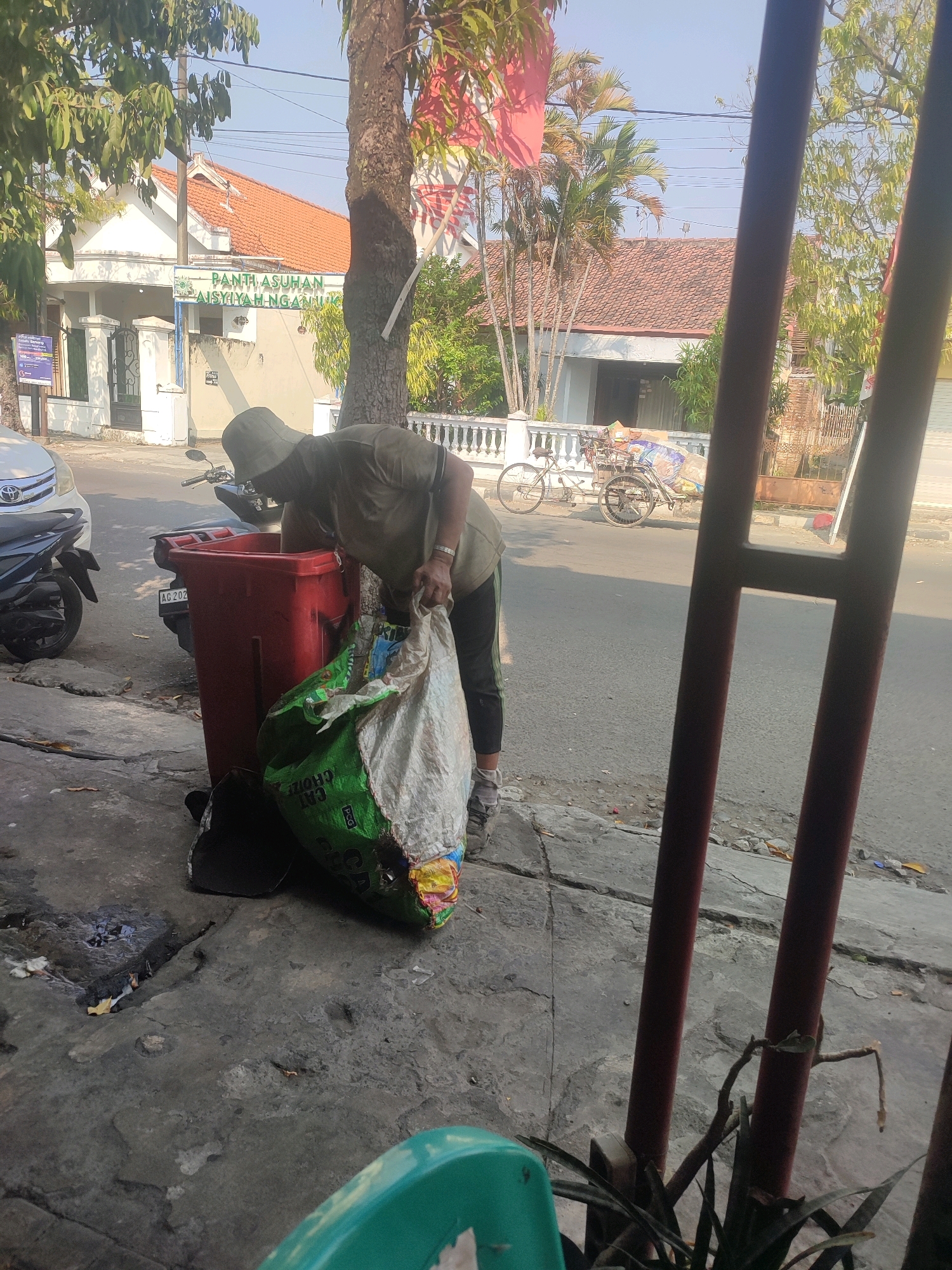 A person wearing a faded olive green short-sleeved shirt, dark pants with a green stripe, a light-colored wide-brimmed hat, and grey athletic shoes is bent over a large red trash bin. The person is holding a large, translucent plastic bag filled with trash, seemingly in the process of disposing of it. The bag is predominantly green with some white and multi-colored sections. Next to the red bin is a black object, possibly another trash bin or a piece of discarded material. The scene appears to be on a sidewalk or roadside in front of a building with an orange tiled roof. The building has white walls and a sign with green lettering that reads "PANTH ASUHAN AISYIYAH NGANJUK," indicating the location is in Nganjuk, Indonesia, and it is likely an orphanage. Further down the street, a bicycle with a cart attached is visible, as are several motorcycles and a white car. The environment is sunny with a clear blue sky. Green foliage from trees and palm trees is present. The foreground shows a concrete sidewalk with cracks and some debris.