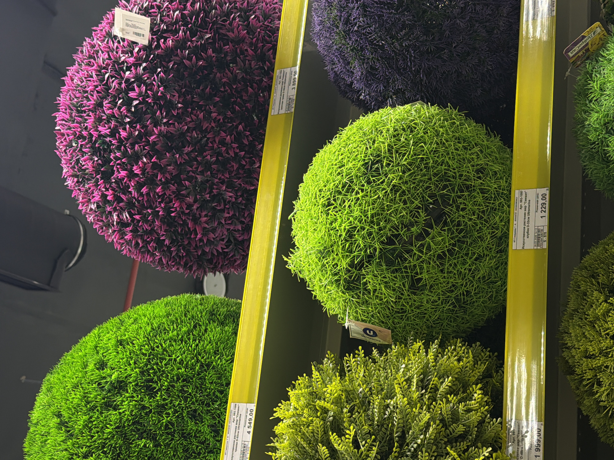 This indoor shot presents a close-up view of a store display featuring decorative plant balls. The balls come in various colors, including pink, dark purple, bright green, lime green, and yellow-green. They appear to be artificial plants arranged into spherical shapes. The display is vertically organized, with yellow poles running along the plants. Price tags are visible near each plant ball, indicating that they are for sale. The image also includes text that is likely Russian. The text mentions "Ultramarine" and "Samshit," possibly referencing the plant types or colors. The price tags display values, suggesting that the photo was taken in a retail store. The text also includes the address of the city of Perm, Russia.