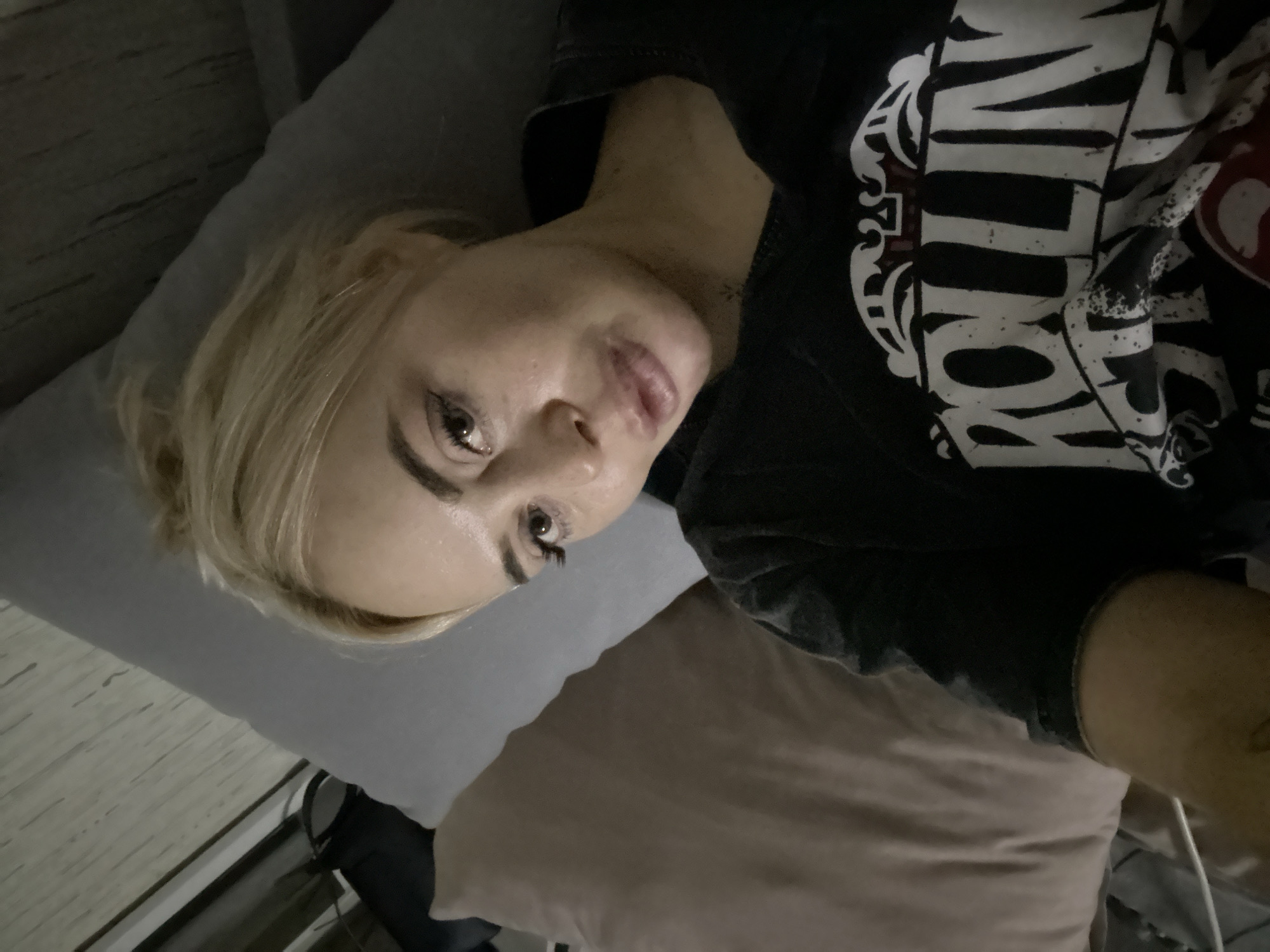 A woman with blonde hair lies on a gray pillow on a light brown sofa. She wears a black t-shirt with white lettering. The location is not specified in the image.