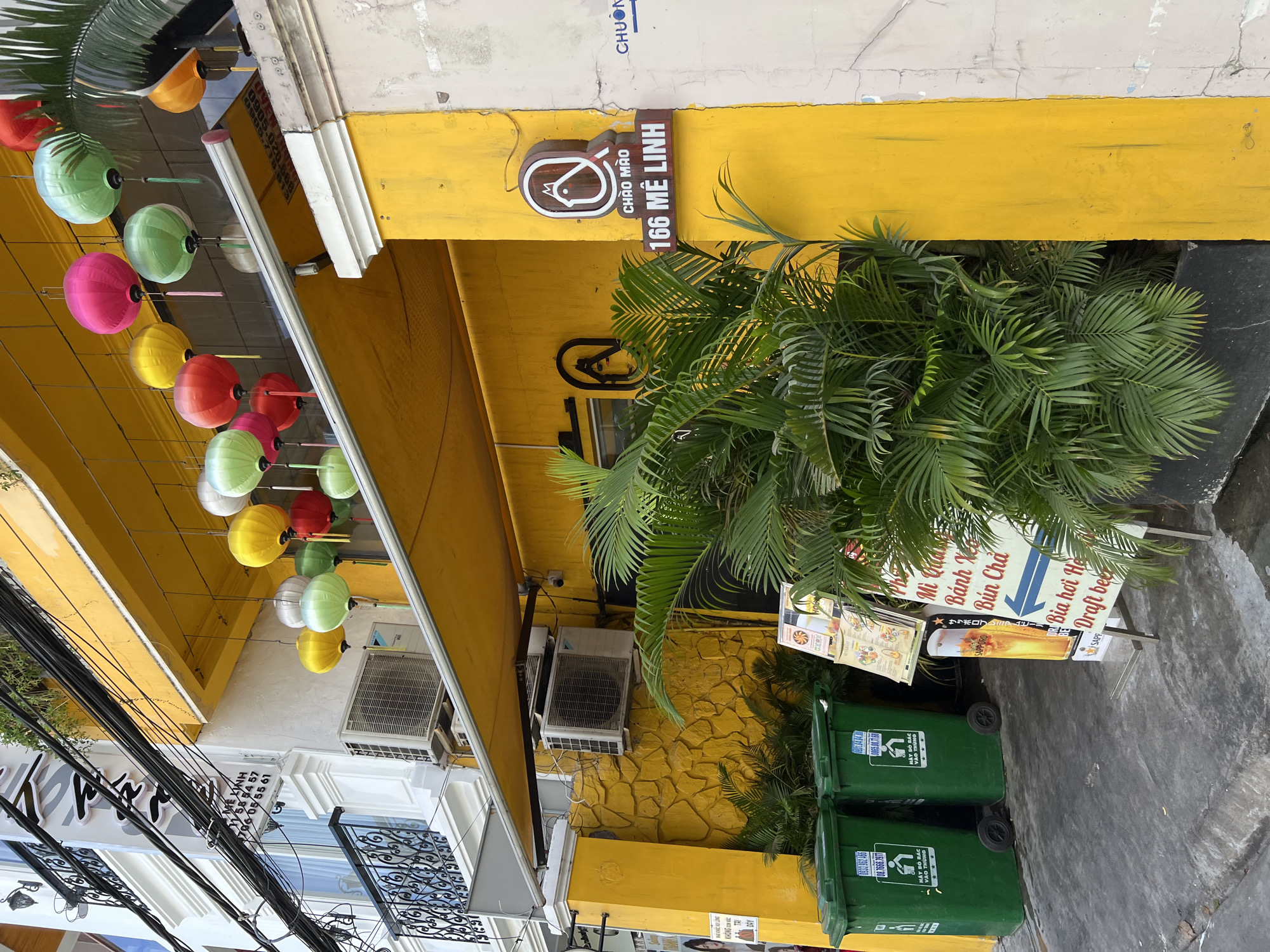 This image depicts the exterior of a brightly painted yellow establishment in Nha Trang, Vietnam, with a focus on its entrance and decorative elements. The building features a yellow awning extending over the entrance, beneath which a collection of colorful, round paper lanterns in red, green, pink, and yellow hang suspended. A lush palm plant is positioned in front of the entrance, partially obscuring it.
To the left of the entrance, two large green trash receptacles with lids are visible, each bearing text in Vietnamese and what appears to be a phone number. Mounted on the wall behind the trash cans is an air conditioning unit. A stone-textured yellow wall forms part of the building's facade.
On the right side of the entrance, a prominent yellow pillar supports the building's structure. Attached to this pillar is a dark brown sign with white lettering that reads "CHAO MAO 166 ME LINH". Above this sign, a circular wooden emblem featuring a stylized chicken or bird logo is mounted. Adjacent to this sign, on the white-colored wall of the neighboring building, a blue arrow points left with the word "CHƯƠNG" written above it.
Additional details include electrical wires running overhead, an air conditioning unit mounted on the upper story of the adjacent white building, and metal railings on balconies of that building. A menu stand is visible near the entrance, displaying various food and drink items, including an image of a beer glass. The ground in front of the establishment is a concrete surface.