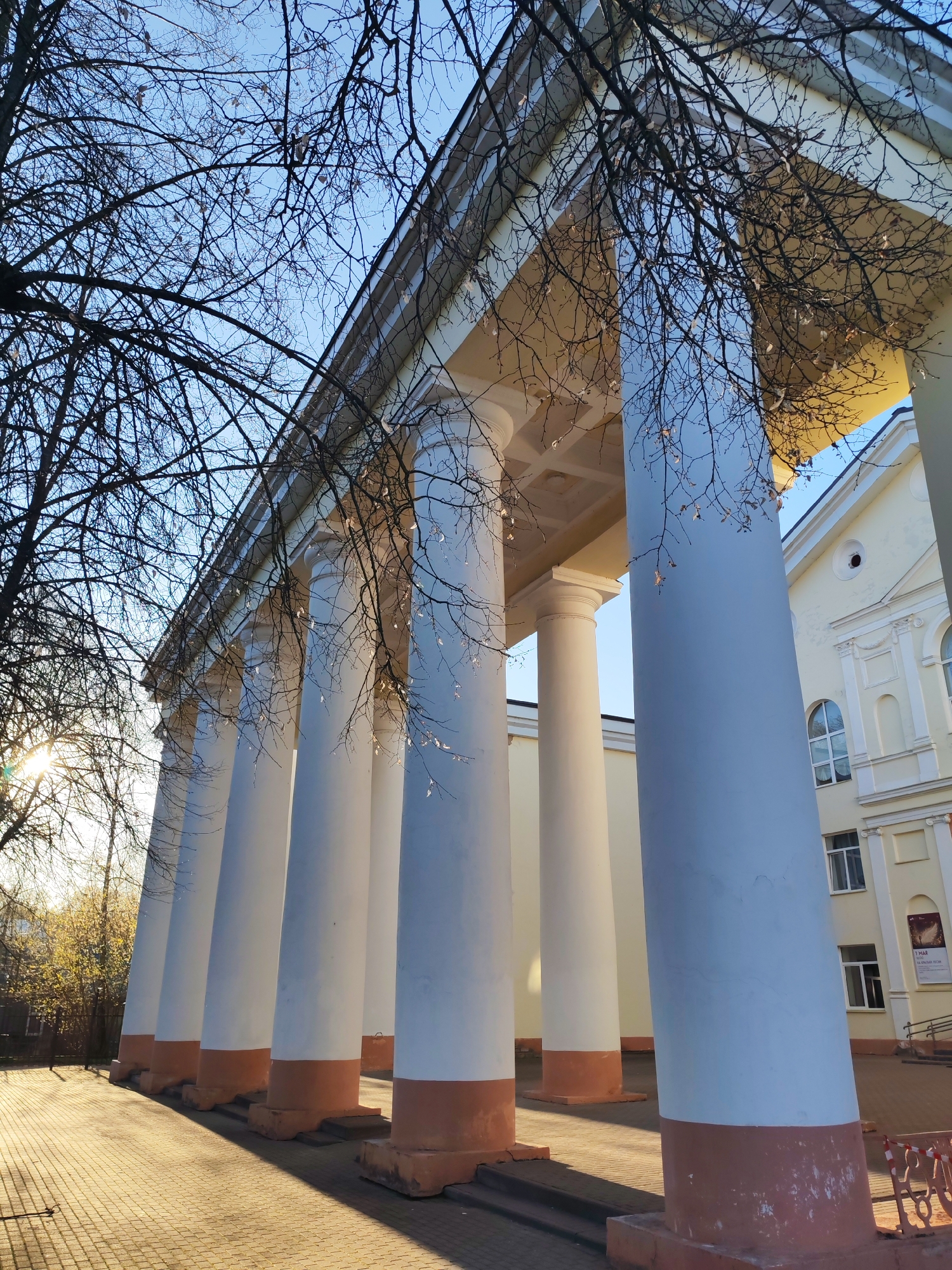 The image depicts a building with a prominent colonnade located in Yakhroma, Russia. The colonnade consists of at least six white columns with a reddish-brown base, supporting a flat roof structure. The building itself is a light yellow color, with arched windows visible on the right side of the frame. Bare tree branches are visible in the foreground and background, partially obscuring the building. The ground is paved with bricks, and long shadows are cast by the columns due to the angle of the sun. The sky is blue, with sunlight filtering through the trees on the left side of the frame.