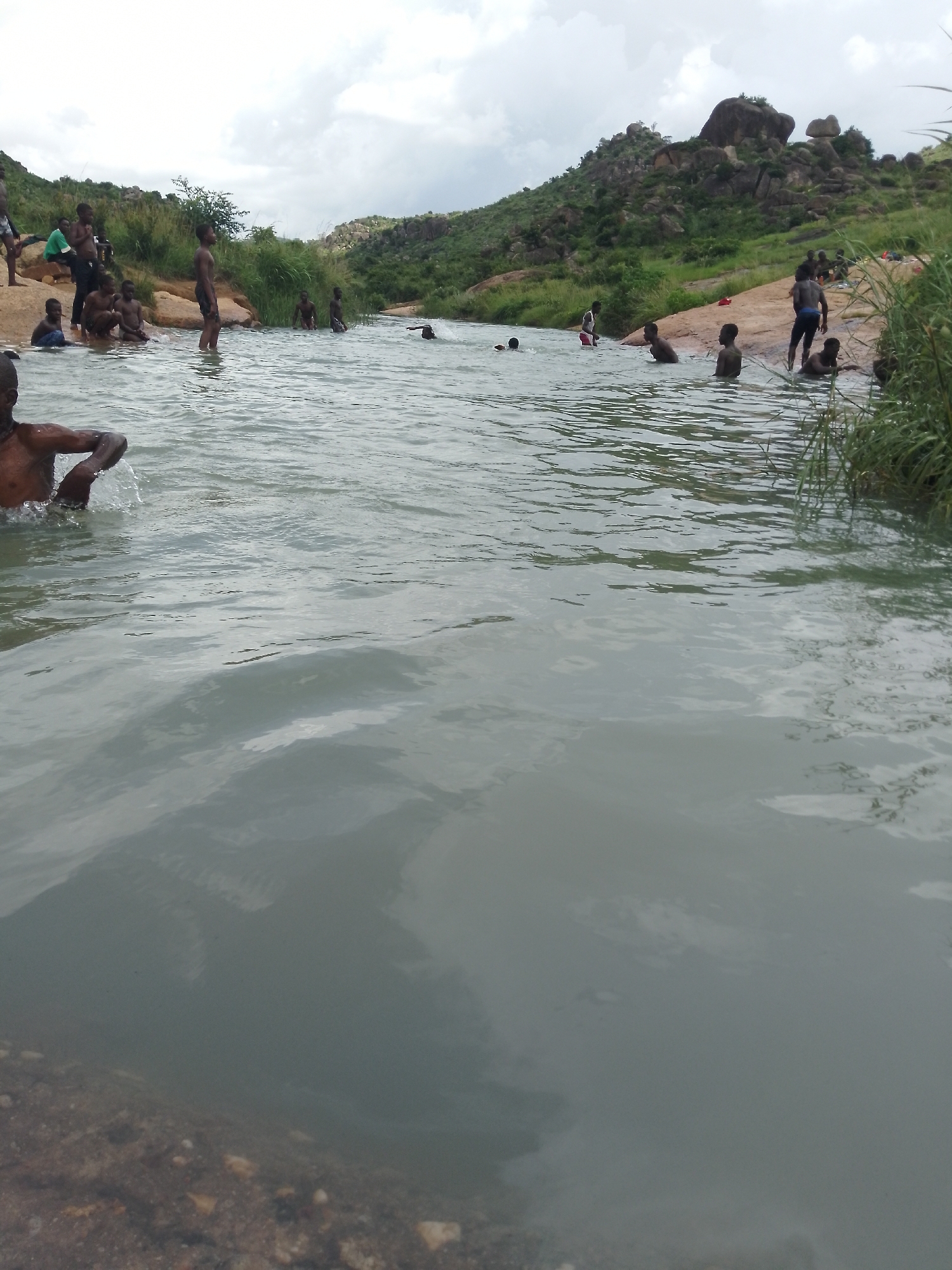 This image captures a scene at the Assop Falls in Jos, Nigeria, where a group of individuals, primarily young men and boys, are swimming and interacting in a river. The foreground features the water with rocky edges, hinting at the natural environment of the falls. In the mid-ground and background, people are visible in various states of engagement with the water. Some are fully submerged, while others are wading or standing near the riverbanks. The environment is characterized by lush green vegetation and rocky hills characteristic of the Jos Plateau. The sky is overcast with clouds, suggesting a possibly humid or rainy day. The overall atmosphere appears relaxed and recreational.