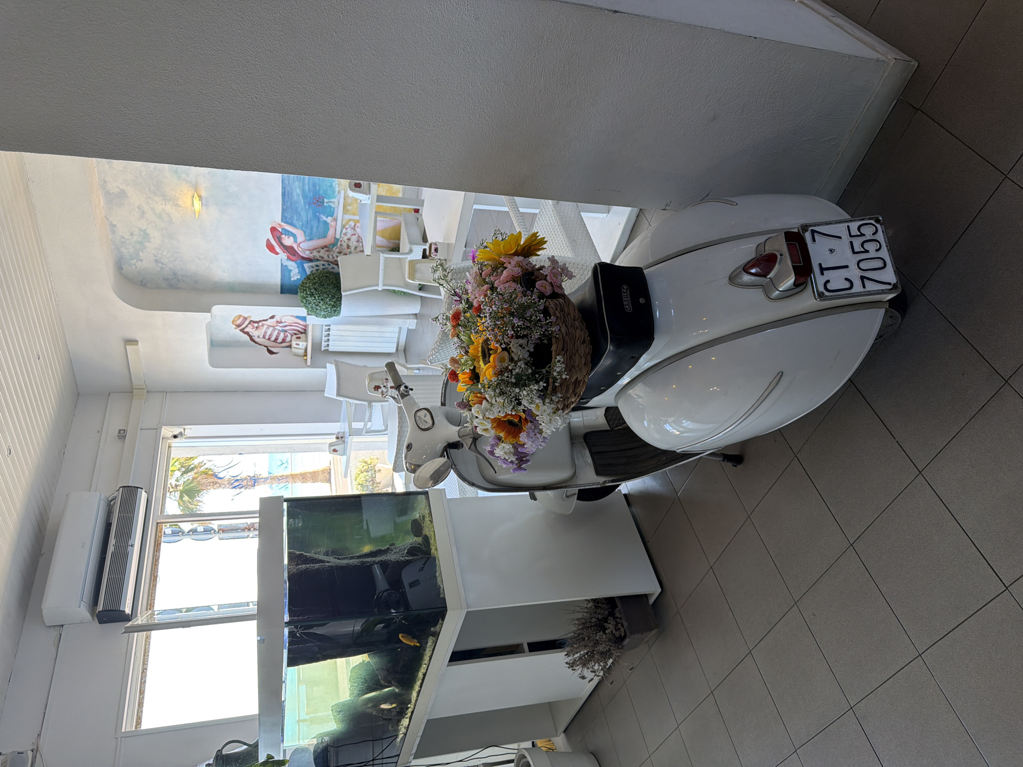 A white Vespa scooter, decorated with a basket of colorful flowers, is parked indoors in what appears to be a cafe or restaurant in Vallecrosia al Mare, Italy. The scooter is positioned in the foreground, facing away from the viewer, with its license plate clearly visible, reading "CT 7 7055". Behind the scooter, a large aquarium filled with fish and aquatic plants sits on a white stand. Further in the background, tables and chairs are set up, suggesting a dining area. A mural depicting figures in beach attire adorns the wall. The room is illuminated by natural light streaming in from a large window that offers a view of palm trees and the sea. An air conditioning unit is mounted on the ceiling. The floor is tiled.