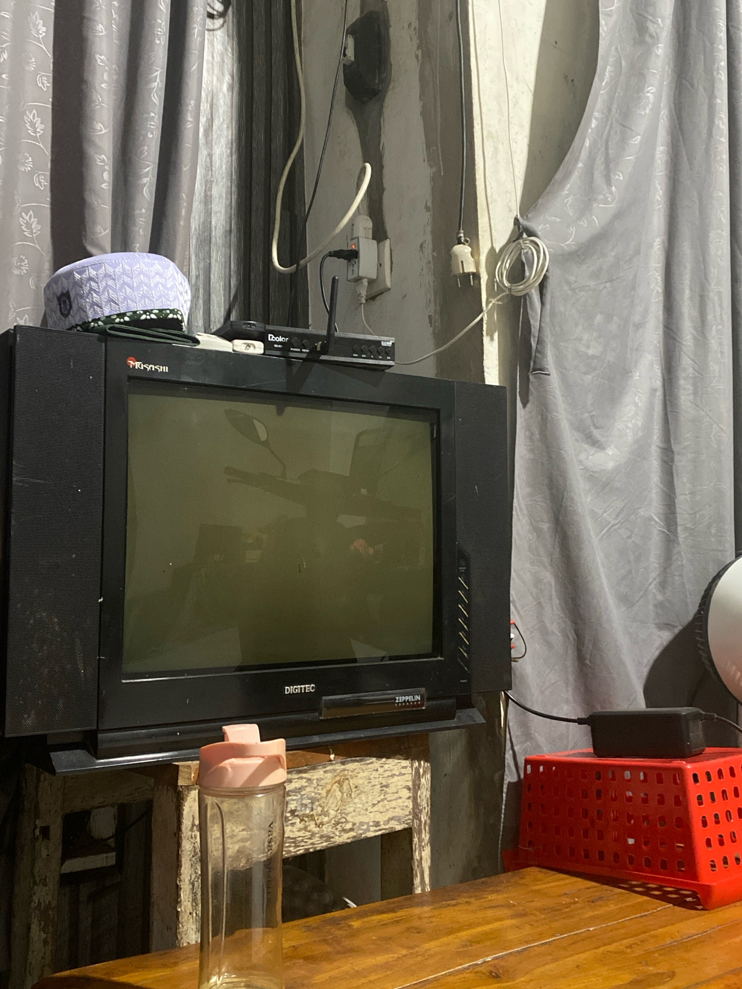 In an indoor setting, likely a living room or bedroom, a vintage black CRT television is positioned on a rustic wooden stand. Above the television, a Muslim prayer cap, white with a geometric pattern and a green and black trim, rests on top of the TV's speaker. Next to the cap, a small black electronic device with "Dcolor" branding is visible. The television screen displays a faint reflection of what appears to be a motorcycle.
To the right of the television, a tall, clear glass bottle with a pink lid is placed on the wooden stand. Further to the right, a bright red plastic basket with a grid pattern sits on the same stand. A black power adapter is placed on top of the red basket. The background is dominated by gray curtains with a subtle floral pattern, drawn to the sides, revealing a textured concrete wall. Electrical cords and a white electrical outlet are visible on the wall. A wooden table is in the foreground, with the edge of the red basket and the glass bottle visible on it. The overall impression is one of a lived-in, slightly cluttered domestic space.