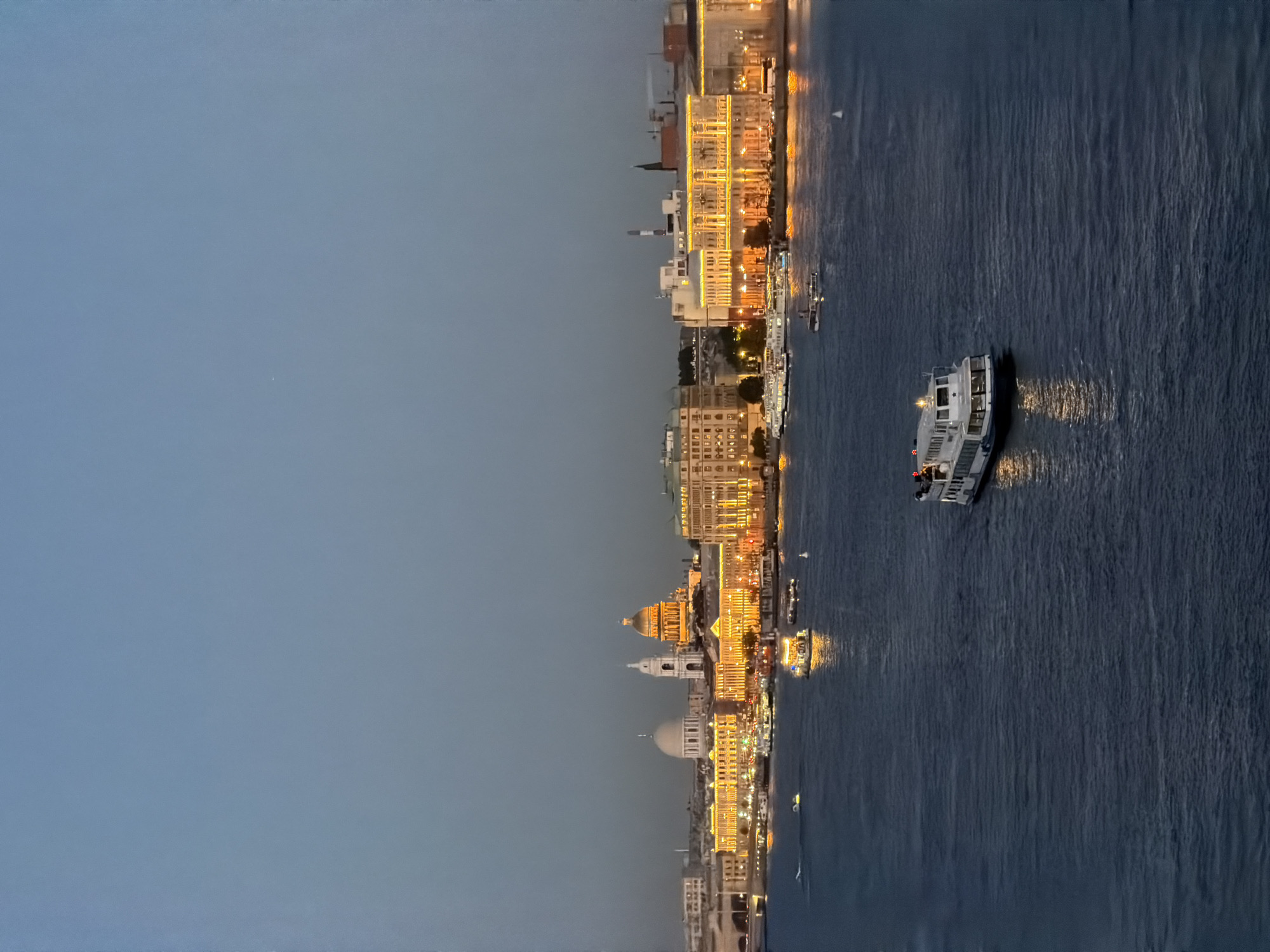 The image depicts the Neva River in Saint Petersburg, Russia, at dusk. A white passenger boat with lit windows is moving across the dark blue water in the foreground. In the background, the illuminated skyline of Saint Petersburg is visible, featuring prominent domed buildings and structures, including what appears to be St. Isaac's Cathedral with its golden dome. Other boats are also present on the river, and the city buildings are brightly lit with yellow lights, creating a warm glow against the twilight sky. The sky is a clear, muted blue, transitioning to a darker hue near the horizon.