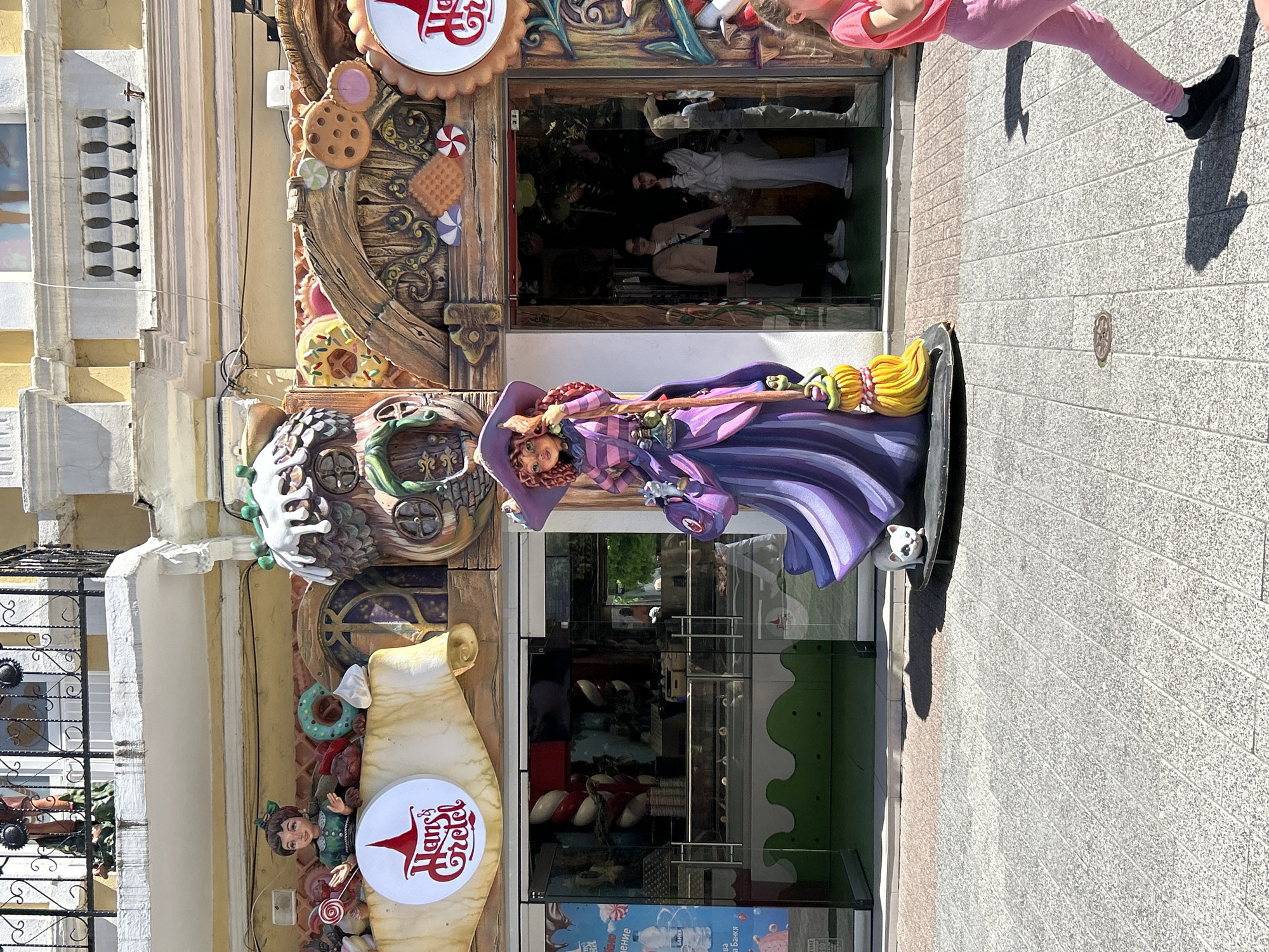 The image depicts the entrance of the Hans & Gretel candy store in Plovdiv, Bulgaria. The storefront is elaborately decorated with sweets-themed ornamentation, including cookies, candies, and gingerbread. A scroll with the "Hans & Gretel" logo is prominently displayed. A statue of a witch with a broom stands to the right of the entrance. The interior of the store, visible through the glass doors, features shelves stocked with candy. Two individuals are visible in the background of the interior. A person wearing pink clothing is partially visible on the right side of the image. The scene is taken during daylight hours. The sidewalk in front of the store is paved.