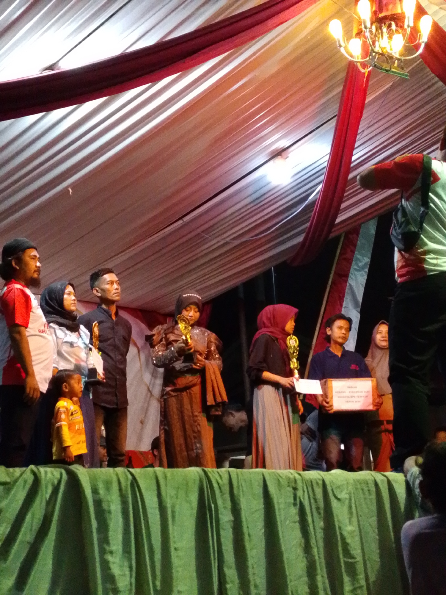 This image depicts an awards ceremony on a stage, possibly outdoors, with a draped canopy overhead. Several individuals, including adults and a child, are present on stage, some holding trophies and certificates. The scene appears to be in Lawatan, Indonesia, given the context. The lighting suggests it is evening or night. The backdrop is a white and red draped fabric, and the stage is covered with a green cloth. A chandelier with multiple bulbs is visible hanging from the canopy. People are dressed in a variety of clothing, including traditional attire and casual wear. Some individuals are smiling, while others appear serious. The overall atmosphere is that of a celebratory event.
