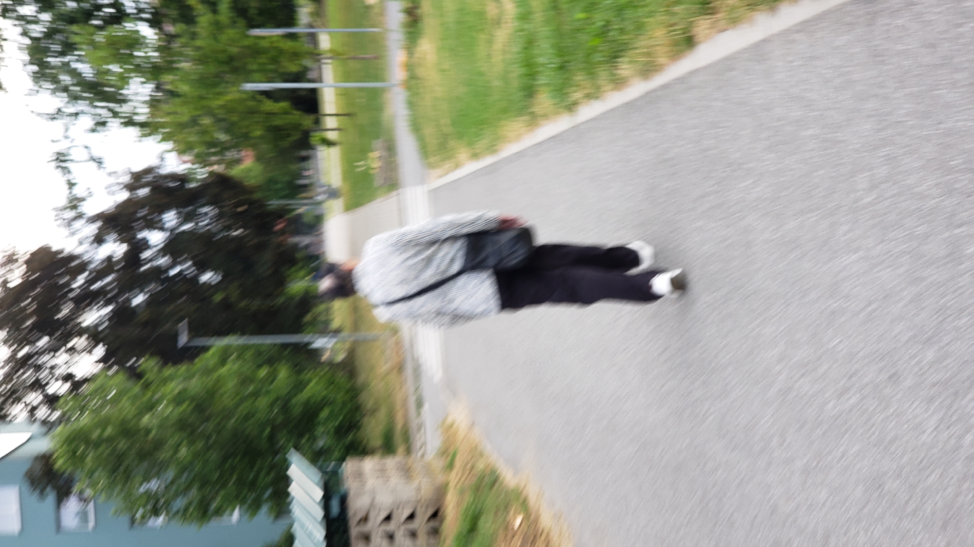 A motion-blurred, medium shot captures a person from behind, walking away from the camera down a paved path in a city park area. The individual wears a horizontally striped, light-colored jacket, dark trousers, and white athletic shoes. A dark shoulder bag is slung across their back. The path is bordered on the right by a grassy area with scattered trees and a few lamp posts. On the left, a light-colored building with visible windows is partially obscured by dense green foliage. A low, concrete structure with several angled openings is also visible on the left. The overall impression is one of a common urban setting, likely a residential area or park in Prešov, Slovakia, with the motion blur suggesting movement and a candid moment.