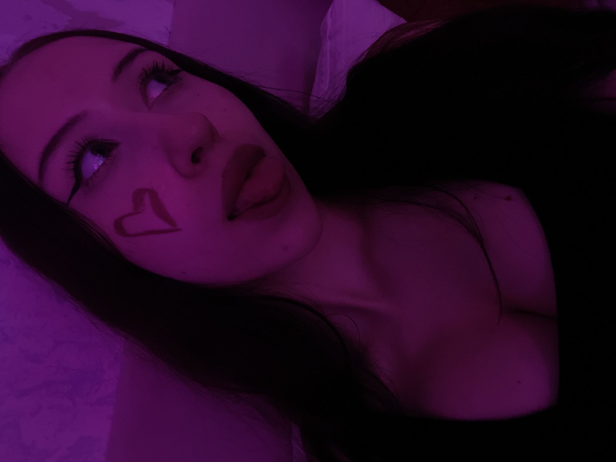 The image shows a close-up, portrait-style photograph of a young woman. She has long, dark hair and is wearing dark clothing. A red heart shape is painted on her left cheek. Her eyes are looking off to the side, and her tongue is sticking out slightly. The lighting in the image is a deep purple hue, casting a dramatic and stylized effect. The background is a textured wall, also bathed in purple light. There is no discernible information about a specific location in Votkinsk, Russia, within the image.