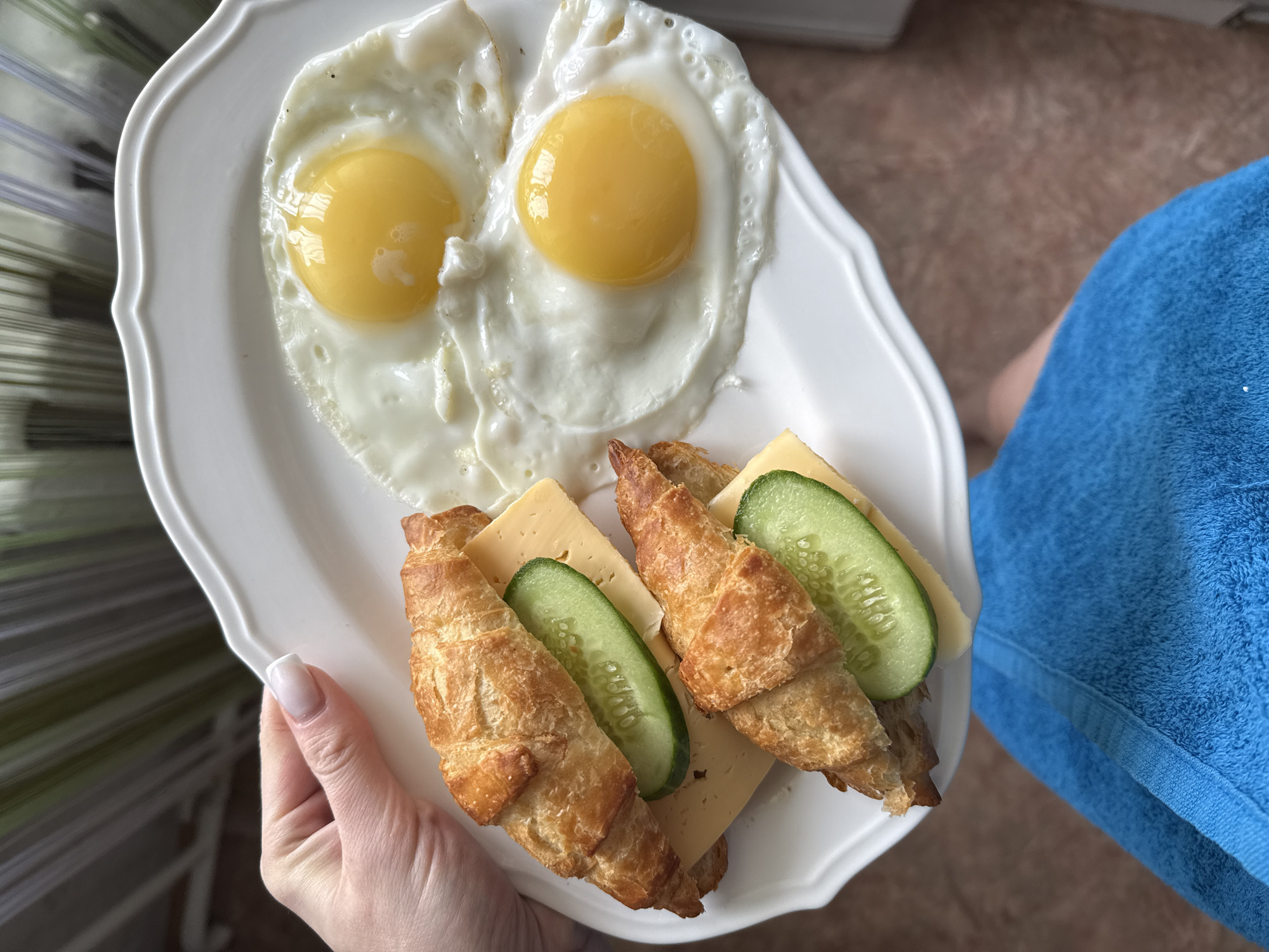 A person holds a white plate with two sunny-side-up eggs and two croissant sandwiches. The sandwiches contain a slice of cheese and two slices of cucumber. The person is wearing a blue garment. The location appears to be Unknown, Unknown.