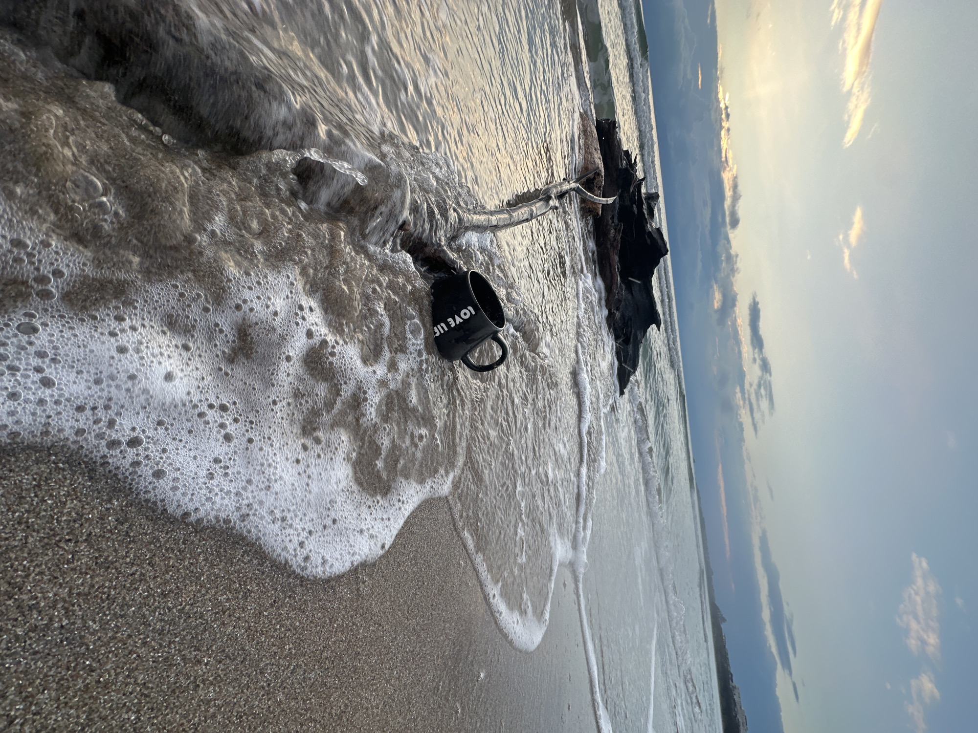 The image shows a black mug with the words "LOVE LIFE" on a sandy beach in Kiten, Bulgaria. The mug is partially submerged in foamy surf washing ashore. A piece of driftwood lies behind the mug, also touched by the water. The background shows a beach meeting the sea and a sky with clouds tinted by the sun. The photo appears to be taken at dawn or dusk. No people are visible.