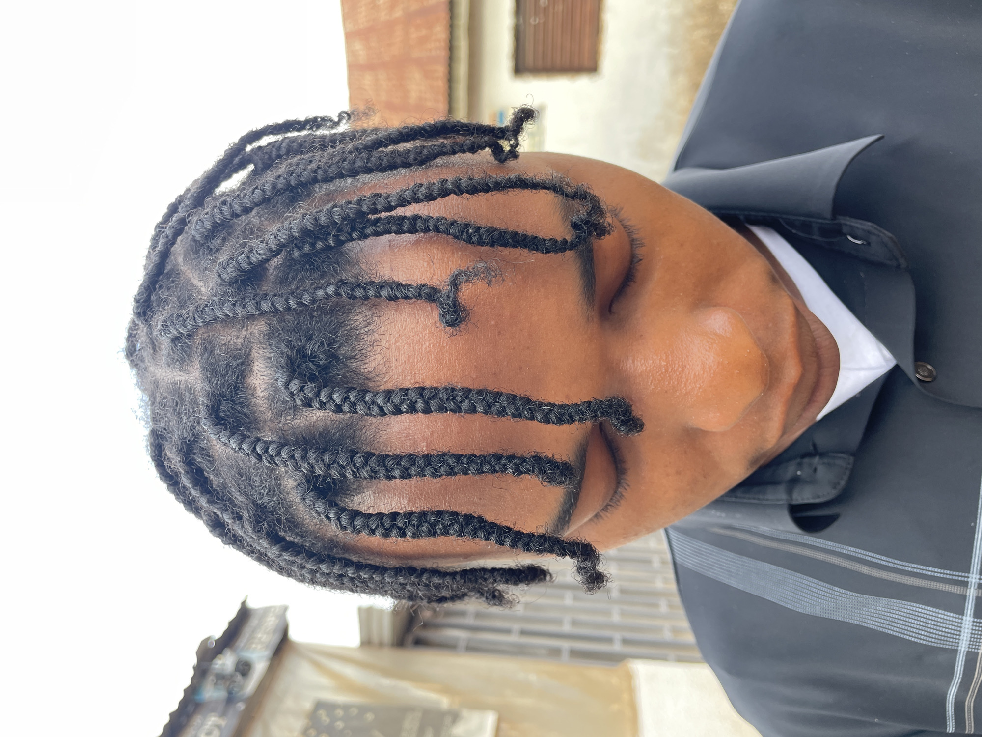 The original image, oriented vertically, depicts a close-up of a person, likely male, with dark skin, in what appears to be an outdoor, daytime setting in Jos, Nigeria. The individual's head is tilted to their right, and their eyes are closed or cast downwards, giving an impression of repose or contemplation.
The most prominent feature is the person's neatly styled hair, which consists of numerous small, dark braids (possibly box braids or twists). These braids are arranged in rows across the scalp, with several strands falling loosely over the forehead and slightly obscuring the closed eyes. The hair texture is visibly curly and coily, typical of natural black hair. Detailed crops reveal the intricate pattern of the braids and the well-defined partings on the scalp.
The person is wearing a black collared shirt or jacket, under which a white undershirt or collar is visible. The black garment has subtle, thin horizontal and vertical grey-white pinstripes or patterns, particularly noticeable on the shoulder and chest area. Buttons are visible on the placket of the black shirt.
The background is largely out of focus, suggesting a shallow depth of field. It shows muted tones of brown, beige, and white, indicating blurred building structures or walls. The lighting is bright and natural, consistent with daylight, illuminating the person's face and hair. No text is visible in the image.