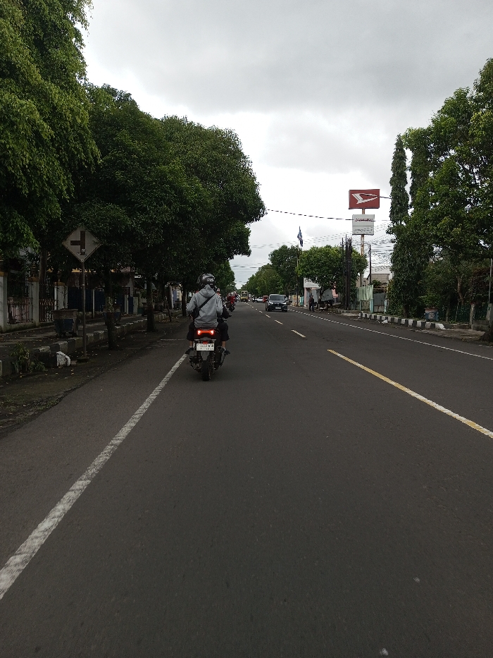 The image depicts a two-lane asphalt road scene, specified as being in the city of Vaatharawattai, Sri Lanka. However, visual elements within the image, specifically a large white sign with a red 'DAIHATSU JOGJA' logo and text, along with a vehicle license plate '1502 WMH' (a format consistent with Indonesia, not Sri Lanka), present a discrepancy regarding this location.
The scene is captured under an overcast sky, showing a dark-colored asphalt road extending into the distance. A solid white line marks the left edge of the nearest lane, and a faded yellow dashed line delineates the center of the road. In the immediate foreground, a dark scooter or motorcycle, viewed from the rear, is traveling away from the camera. It has two illuminated red taillights and a license plate reading '1502 WMH'. Two individuals are on the motorcycle: the driver wears a light-grey hooded sweatshirt and a light-colored helmet, while a passenger sits behind, partially obscured in dark clothing.
On the left side of the road, a sidewalk with a grey curb borders a row of dense green trees. Amidst these trees, a white diamond-shaped road sign with a black T-junction symbol (indicating a main road with a right-side junction) is visible. Behind the trees, structures with fences are partially discernible.
On the right side, parallel to the road, another line of green trees stands. Above these trees, a prominent white billboard displays a red 'DAIHATSU' logo and black 'DAIHATSU JOGJA MANDIRI' text. Utility poles and overhead wires are also present. In the background, additional vehicles, including a dark sedan and possibly a yellow bus, are visible on the road, heading towards the viewer. The overall environment suggests an urban or semi-urban setting.