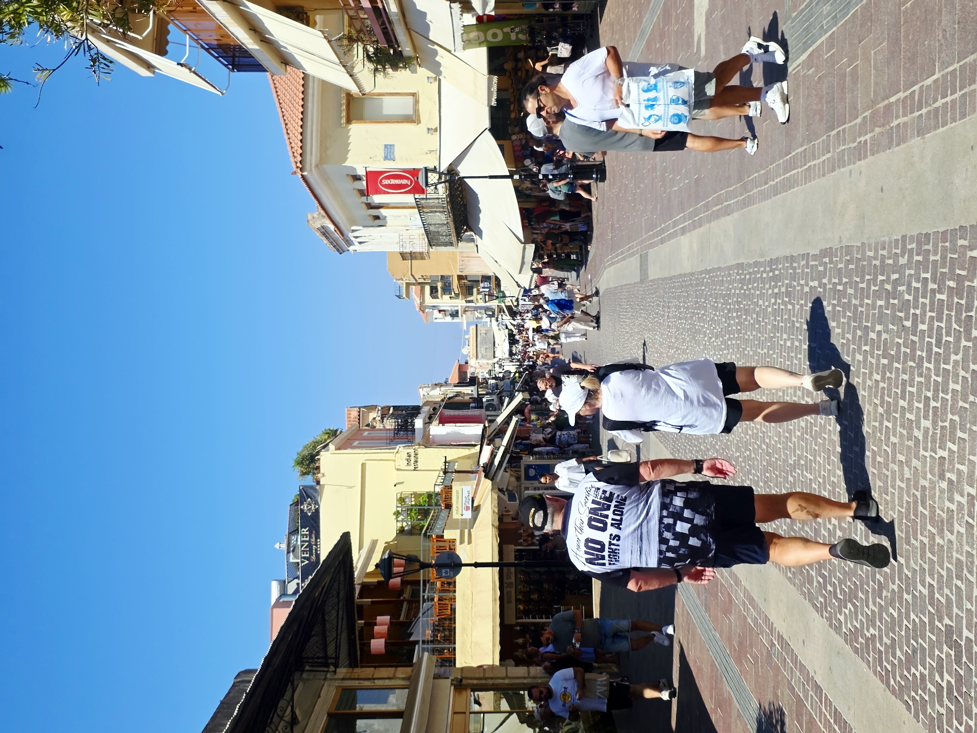 The image depicts a sunny day in the city of Chania, Greece, specifically along a pedestrian street lined with shops and restaurants. In the foreground, two individuals are walking away from the camera. The person on the left wears a black and white patterned t-shirt with the text "NO ONE FIGHTS ALONE" and dark shorts. The person on the right wears a white t-shirt and black shorts. The street is paved with brick and has a concrete central strip. Shops with awnings and balconies are visible on both sides of the street, with signage indicating businesses such as "VENER Luxury Suites" and "Indian restaurant." Several other people are scattered throughout the scene, walking in both directions. The sky is clear blue, indicating good weather.