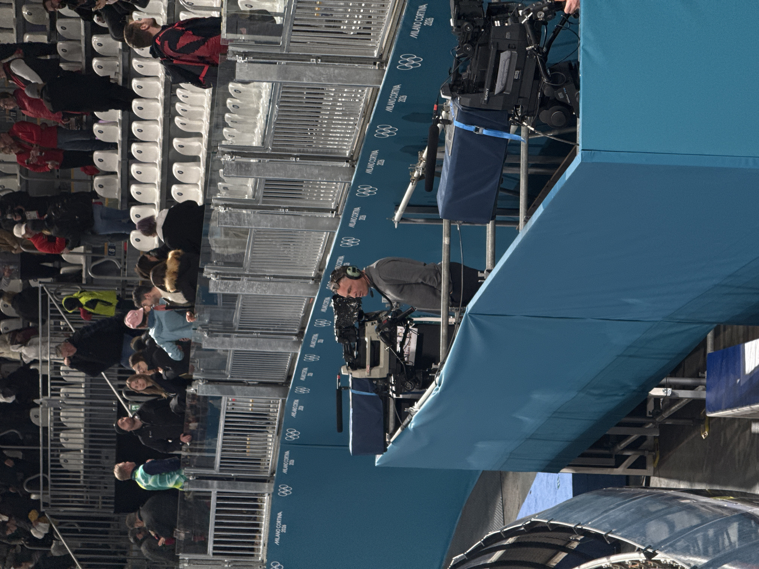 A cameraman with headphones operates a large Sony broadcast camera at a sporting event. The camera is positioned on a platform draped with blue fabric displaying the "Milano Cortina 2026" logo and Olympic rings. Behind the platform are stadium seats, some occupied by spectators. The scene appears to be outdoors, possibly during a competition. The setting suggests an official broadcast setup at a sports venue, likely for the 2026 Winter Olympics.