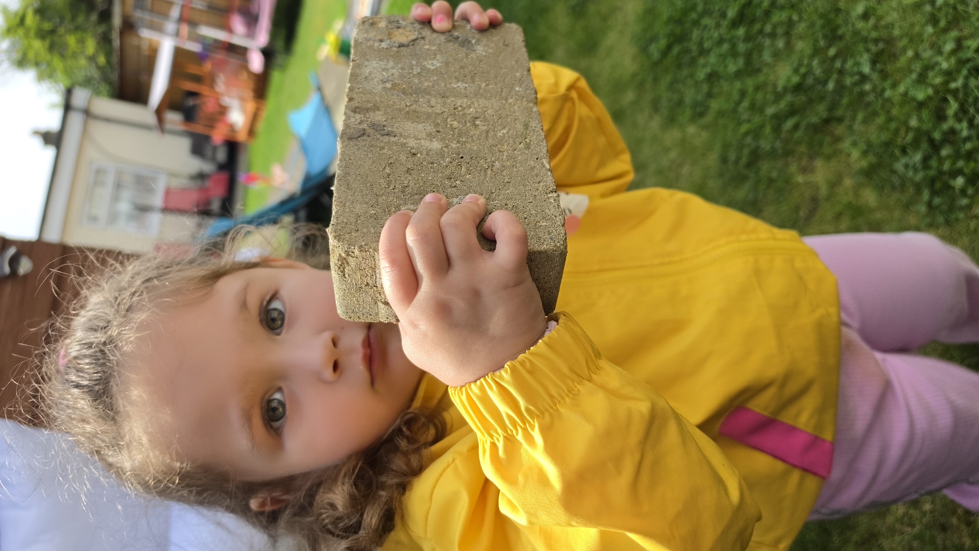 A young child is lying down in a grassy area, holding a grey concrete brick up in front of their face. The child has fair skin, light brown, curly hair and is wearing a yellow raincoat with a pink accent. They are also wearing pink trousers. Their hands are holding the brick in a way that partially obscures their face. The background includes green grass, a playhouse and a blurred building. The location is likely a residential garden. The specific location in London, United Kingdom, cannot be determined from the image alone.