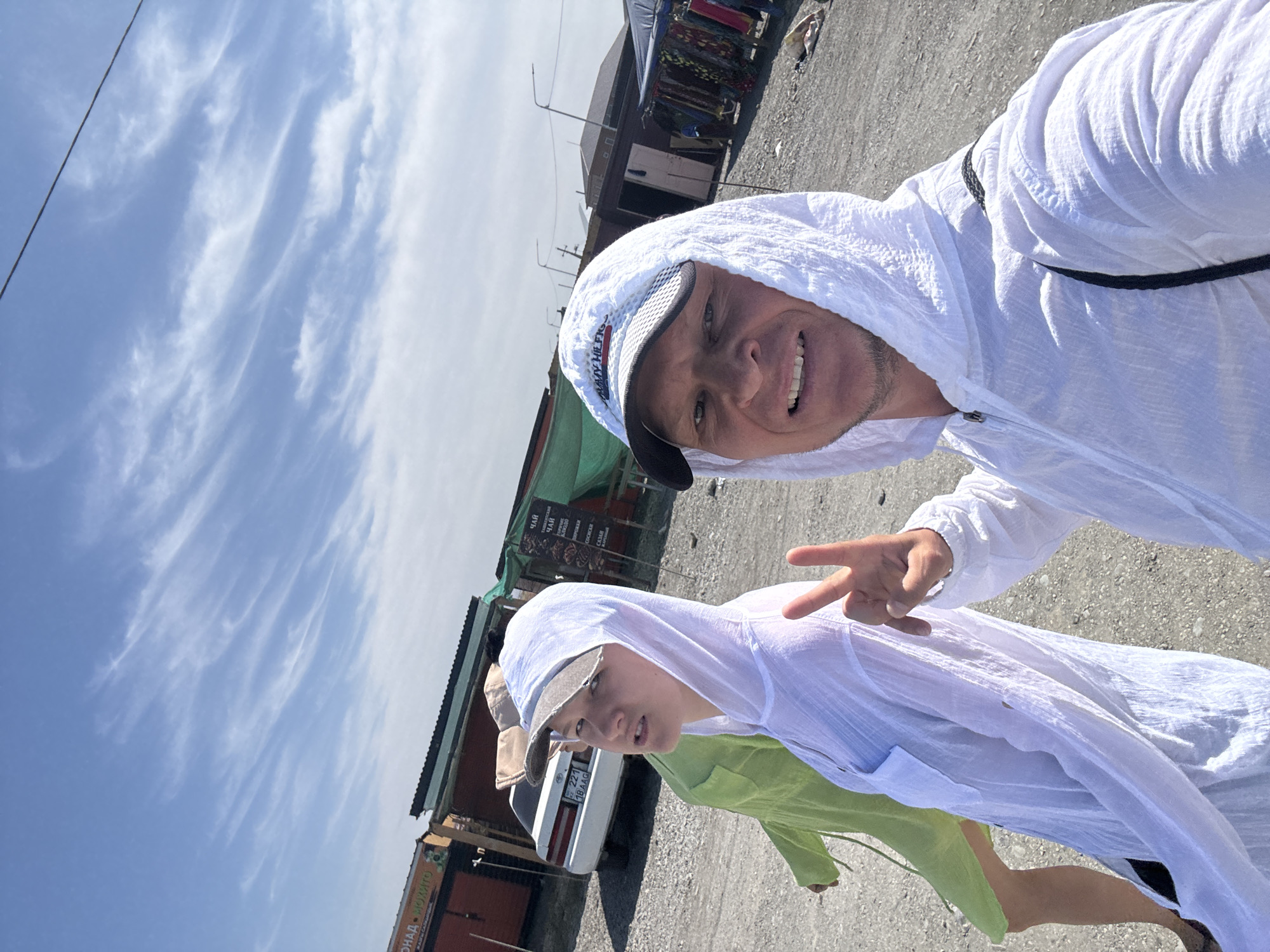 The image shows two individuals walking outdoors in an area that appears to be a market or roadside stalls. The person in the foreground on the right is a man wearing a white hooded jacket and a white and black baseball cap, making a peace sign with his right hand. The person in the middleground on the left is a younger individual wearing a white hooded jacket and a baseball cap, looking towards the camera. Behind them, a person in a light green garment is visible. Several stalls with awnings are set up, some displaying goods like clothing. A white car is parked in the background. The ground is a gravelly surface. The sky is blue with wispy white clouds. The overall impression is of a sunny day in a somewhat rustic, open-air commercial setting. Based on the context provided, this scene is located in the city of Zharbulak, Kazakhstan.