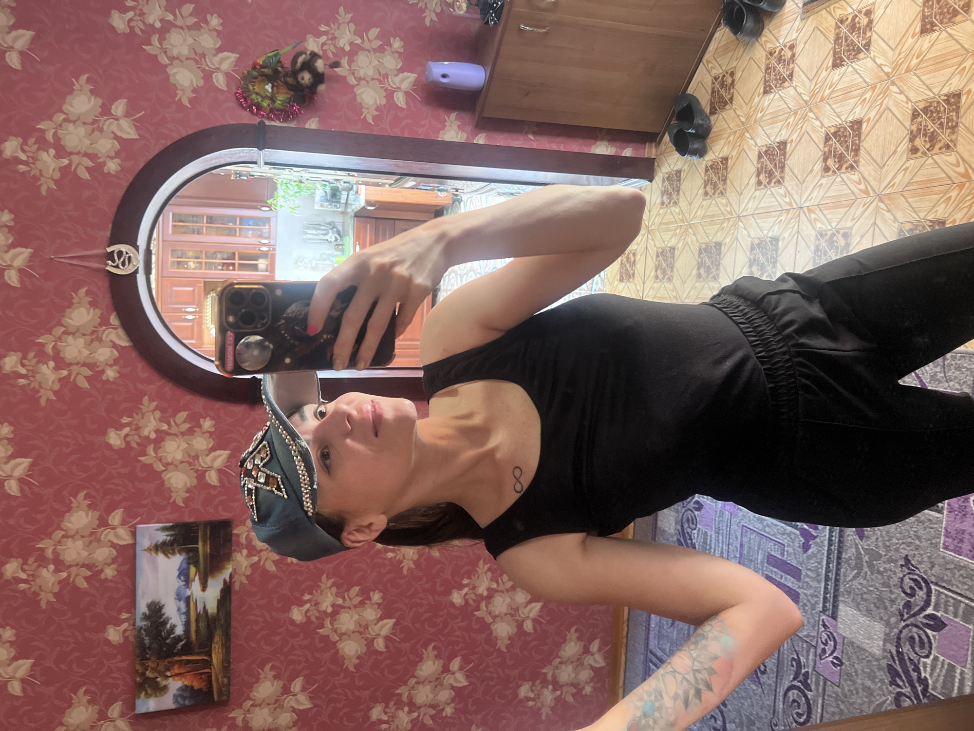A woman takes a selfie in a room with pink floral wallpaper. She is wearing a black tank top, black sweatpants, and a teal baseball cap adorned with rhinestones and a star design. A tattoo is visible on her left forearm, and an infinity symbol tattoo is on her chest. She is holding a smartphone to take the picture. In the background, through an arched doorway, a kitchen area with wooden cabinets is visible. To the left of the doorway, a landscape painting hangs on the wall. On the floor, black slip-on shoes are present. The overall scene suggests an indoor domestic setting, possibly in Krasnoe-na-Volge, Russia, given the context.