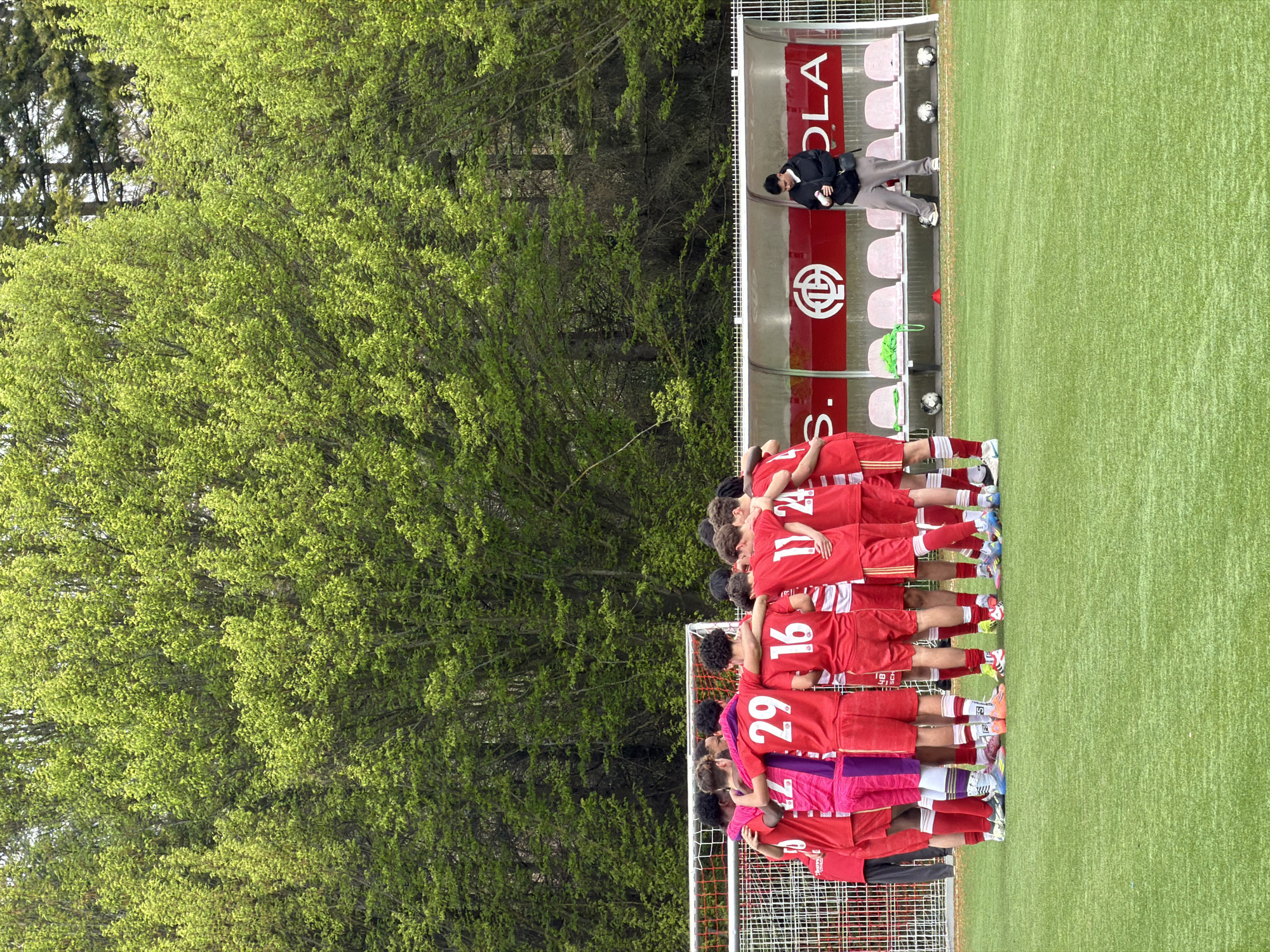 The image shows a soccer team huddling together in a group on a green field, likely before or after a game. Most players are wearing red jerseys with white numbers on the back, except one player who is wearing a magenta jersey with purple shorts. The jerseys are numbered 29, 16, 11, 24, and others are visible. A team manager dressed in black is standing behind the covered player benches. Behind the team is a forest and the covered player benches, which are red and white and say "S. COLA" and contain a red cross logo, which implies that this is a team in Esch-sur-Alzette, Luxembourg.