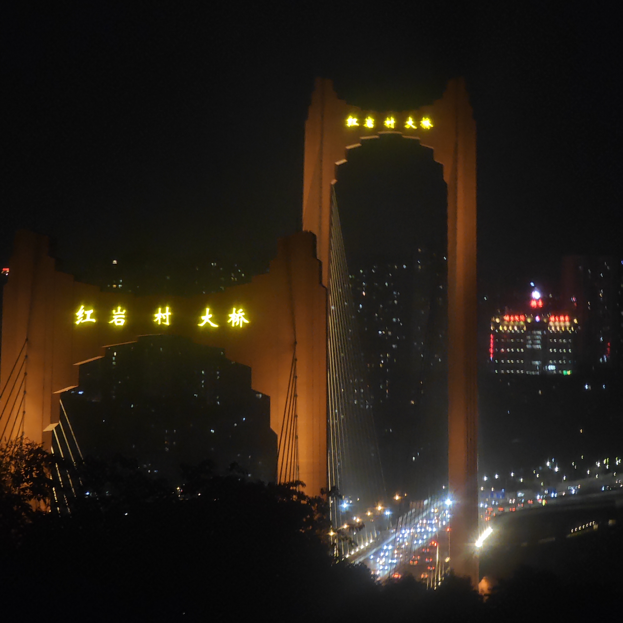 The image captures the Hongyancun Bridge in Yuzhong District, China, at night. The bridge's illuminated towers are prominent, displaying Chinese characters in bright yellow lights. In the background, city lights twinkle against the dark sky, including a lit-up building with red and yellow lights. At the bridge's base, traffic moves along the roadway, creating streaks of light. Silhouetted trees and foliage are in the foreground, adding depth to the scene.