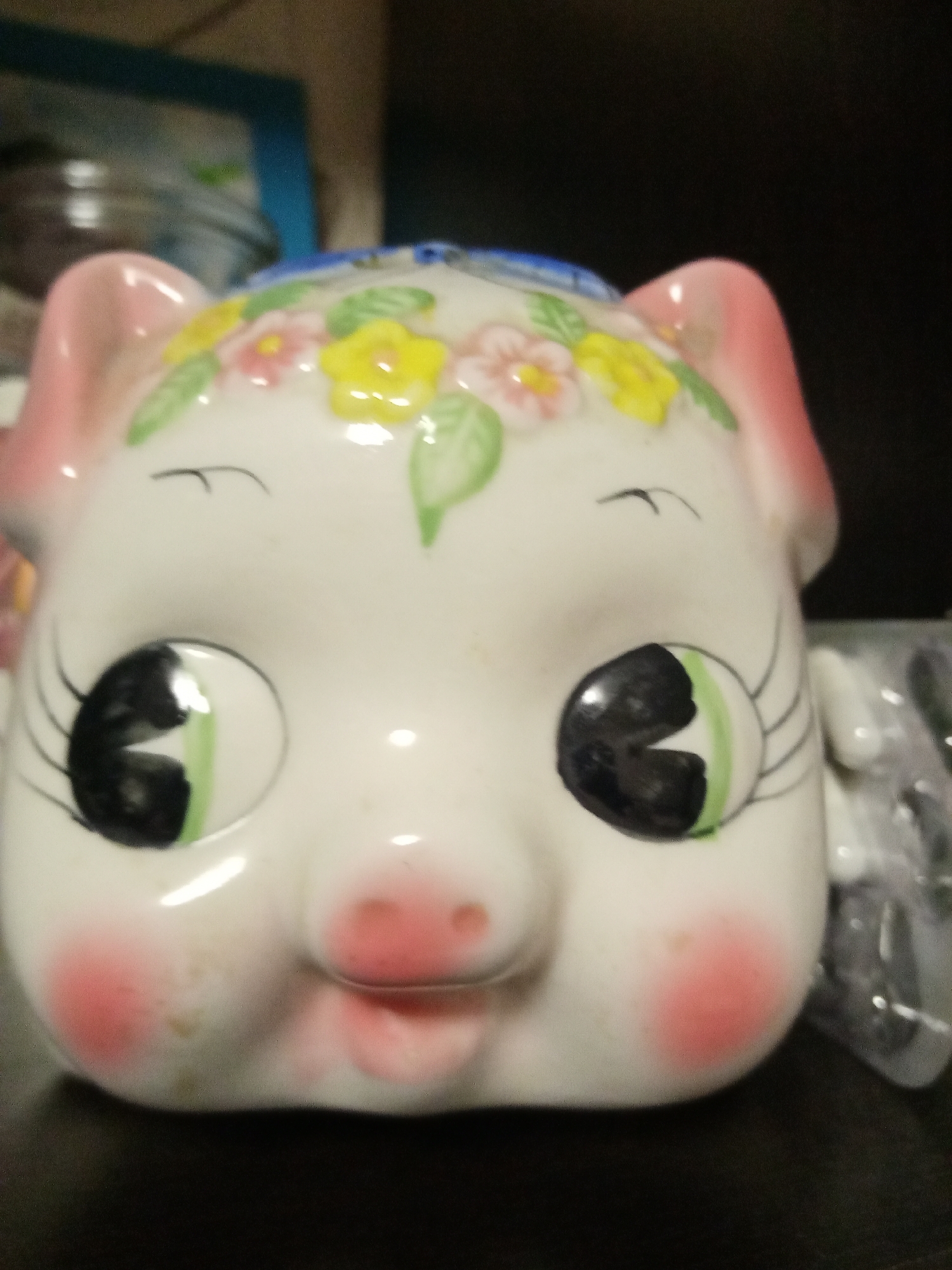 A close-up photograph shows a ceramic piggy bank with a pig face. The pig is white with pink ears and cheeks, and it has drawn-on eyelashes. The piggy bank is decorated with yellow and pink flowers and green leaves on its head. The location and city of Elektrostal, Russia are not discernible from the image.