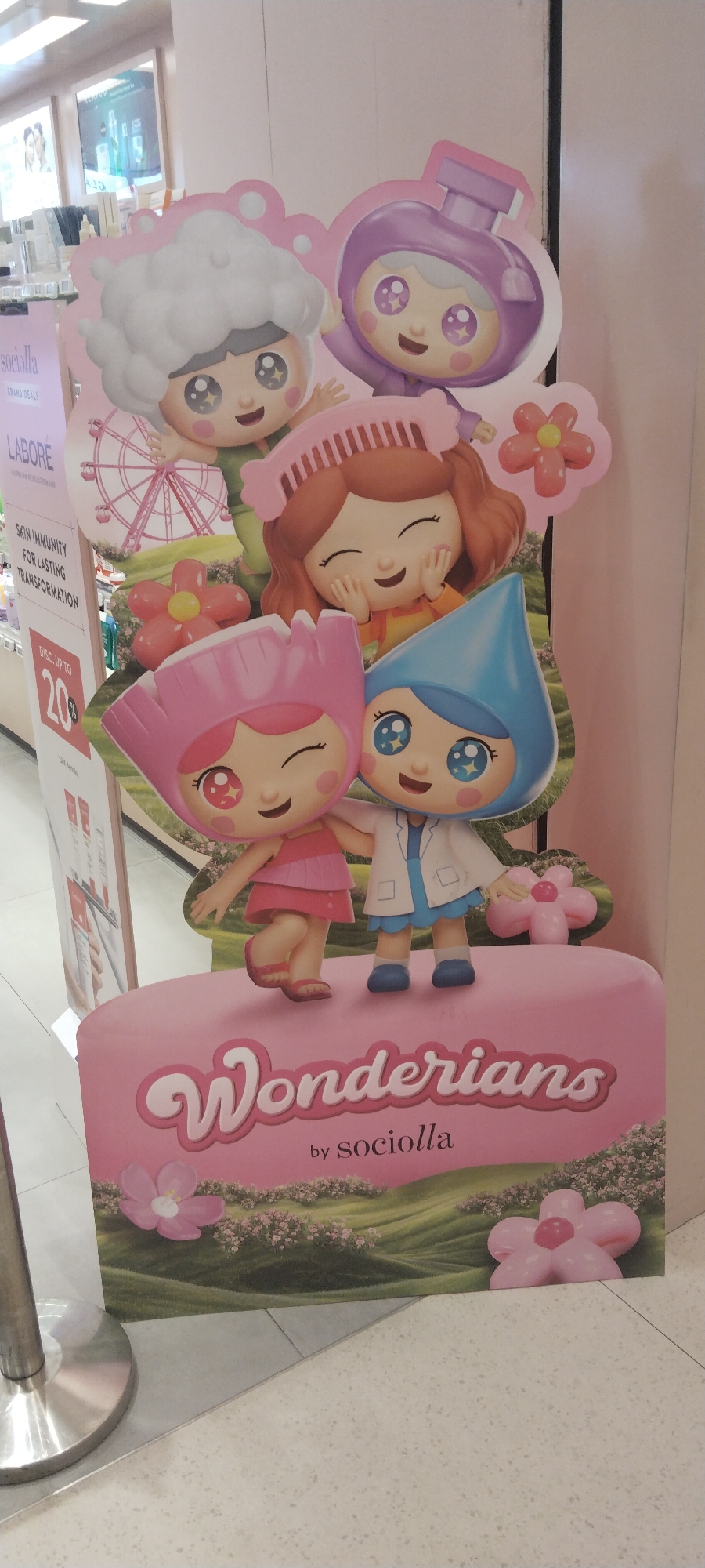 A life-sized cardboard cutout display featuring four cartoon characters is positioned in an indoor retail environment. The display is pink and features a grassy, hilly background with scattered pink flowers. At the bottom of the display, the words "Wonderians by Sociolla" are written in stylized pink text.
The characters are:
- On the top left, a character with a large white, cloud-like hairstyle, a green shirt, and a Ferris wheel in the background.
- To the right of the first character, a character wearing a purple outfit resembling a perfume bottle, with a pink flower to its right.
- Below the first character, a character with a pink helmet, a pink dress, and holding the hand of the character to its right.
- To the right of the character in the pink dress, a character wearing a blue, teardrop-shaped hat and a white lab coat, with a pink flower to its right.
To the left of the display, there is a vertical retail signage stand with text that includes "sociolla," "BRAND DEALS," and "LABORÉ." Below this text, there is information about "SKIN IMMUNITY FOR LASTING TRANSFORMATION" and a "DISC. UP TO 20%" sale.
The floor is tiled with a light-colored, speckled pattern. A metal stanchion with a brushed metal finish is visible on the far left of the image. In the background, out of focus, are shelves stocked with various retail products, suggesting the location is a store, likely a beauty or skincare store given the branding.