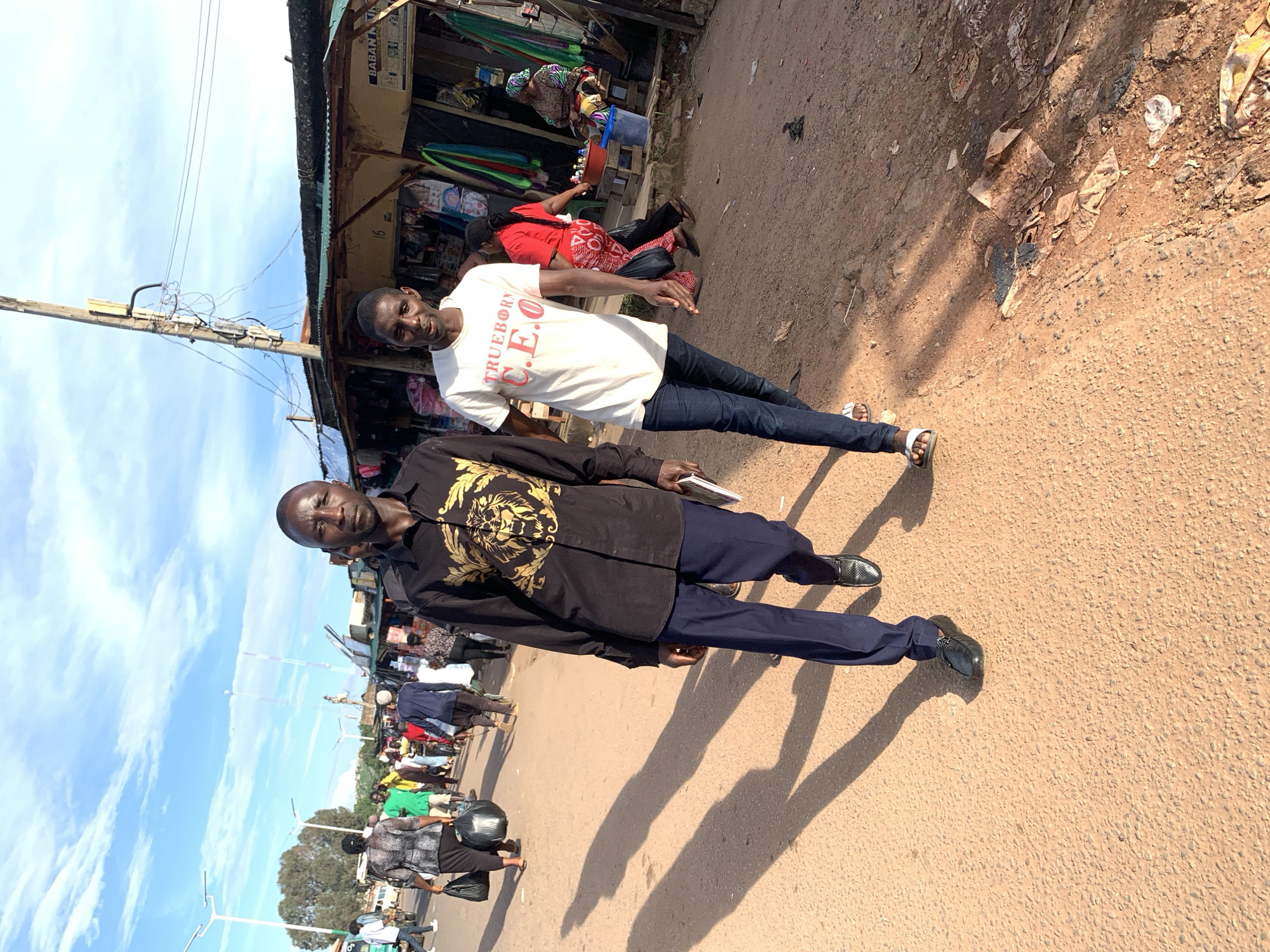 In an outdoor market scene in Jos, Nigeria, two men are walking towards the camera on a dusty, reddish-brown path. The man on the left wears a black shirt with a prominent gold lion design and dark blue trousers, and he carries a book in his right hand. The man on the right is dressed in a white t-shirt that reads "TRUEBORN C.E.O." in red lettering and blue jeans, and he is wearing white flip-flops. Behind them, the market is lined with various stalls constructed from corrugated metal and wood, displaying goods. Several other people are visible in the background, some carrying shopping bags. The sky above is blue with scattered white clouds, and utility poles with electrical wires stretch across the frame. The lighting suggests it is daytime, possibly late afternoon, casting long shadows from the figures onto the ground.