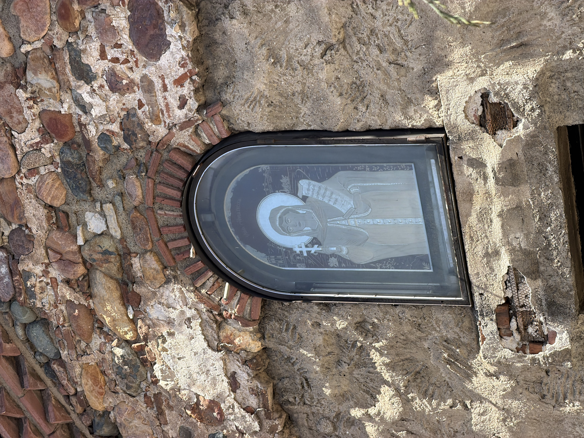 The image shows an icon of a saint, possibly the Virgin Mary, etched on glass and set into a wall made of stone and brick. The wall has an arched opening built with small red bricks, and the glass with the icon is set within this arched opening. The wall itself appears to be made of larger stones and mortar. To the right of the glass frame, the wall shows signs of damage, revealing the underlying structure, including what looks like mesh and possibly wood. The image is taken at an angle, likely with a mobile phone.
Without further context, it's not possible to confirm the exact location of this image in Sofia, Bulgaria. However, given that the image contains a religious icon embedded within stonework, the scene could potentially be located at one of the historic churches or monasteries in or near Sofia, where such religious iconography is common.