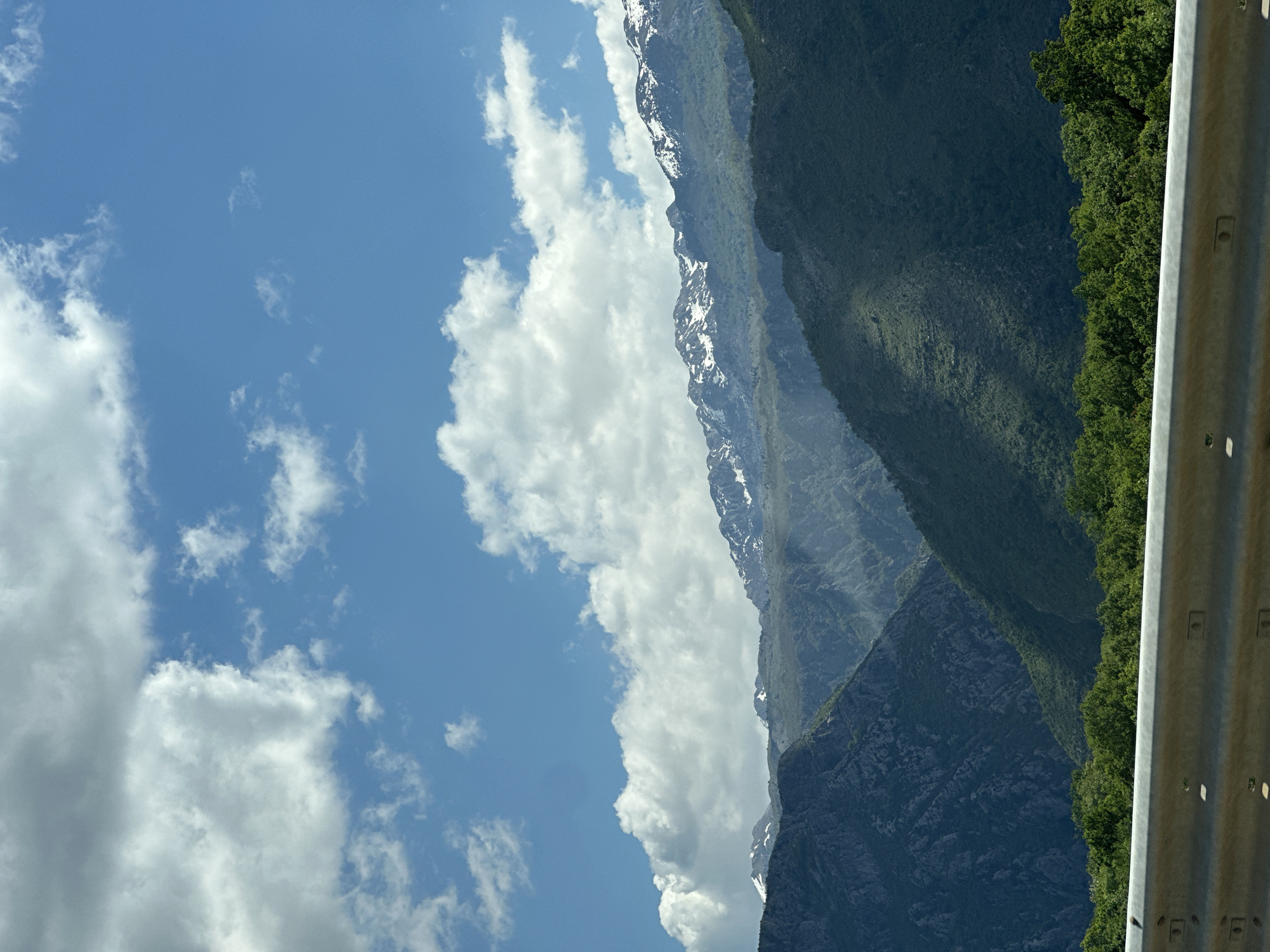 The image shows a view from a roadside in Klopot, Montenegro, featuring a mountain range under a partly cloudy blue sky. Some of the higher peaks have patches of snow. Dense, dark green vegetation covers the lower slopes of the mountains. A grey metal guardrail runs along the right edge of the frame.