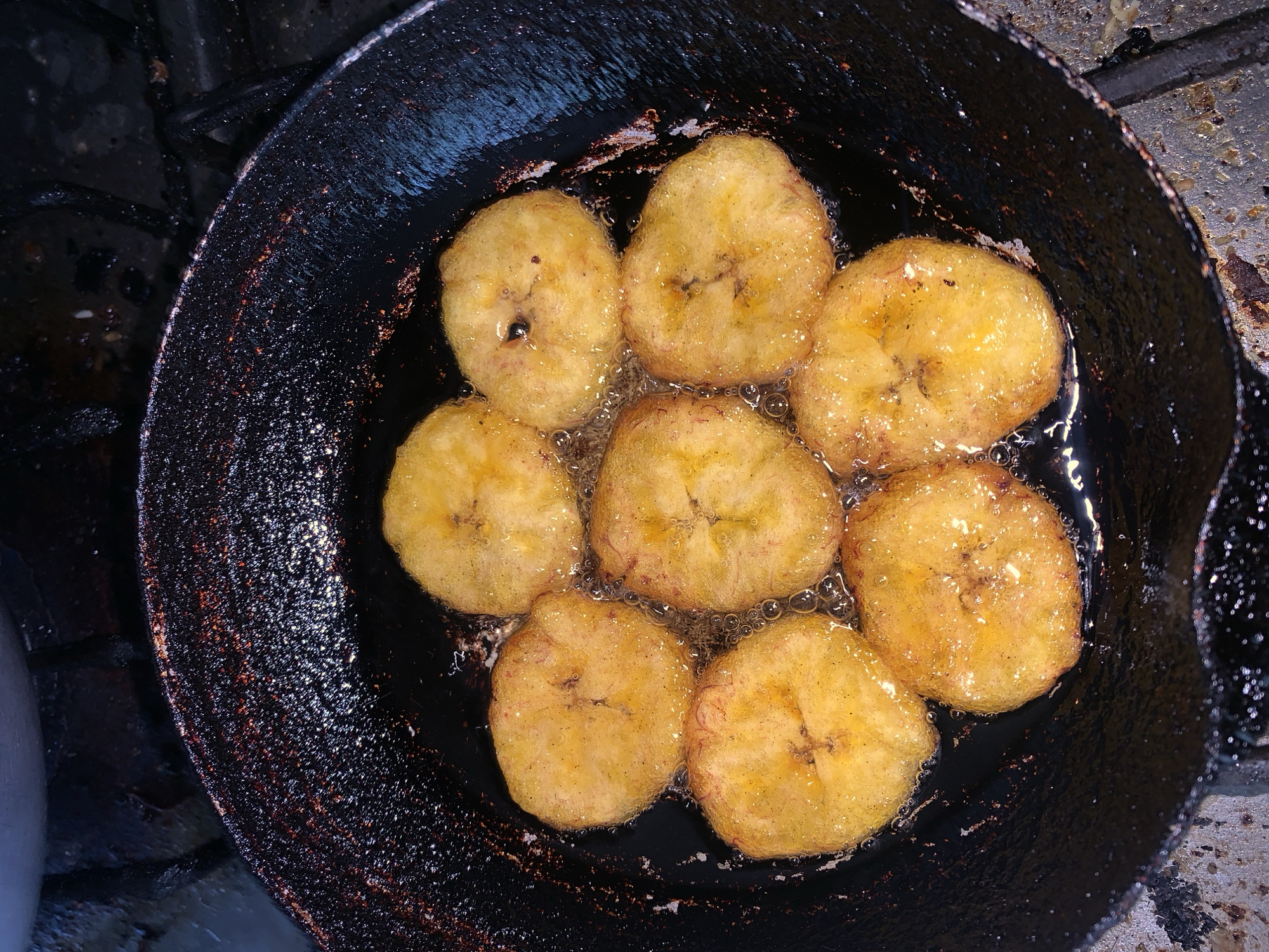 An overhead, close-up shot captures eight circular slices of plantains or ripe bananas frying in a dark, well-used pan. The slices are golden-yellow to light orange, with some showing slightly darker, caramelized spots and faint internal markings from the fruit's core. They are submerged in bubbling, clear oil, indicating active cooking. The hot oil glistens and reflects light, creating numerous small, bright bubbles around the food.
The pan itself appears to be a dark metal, possibly cast iron, with a textured surface and visible signs of use, including grease splatter and wear around its rim. The edges of the pan are dark brown and black, contrasting with the vibrant color of the cooking food. The background outside the pan is dark and indistinct, suggesting a stove top or a dim kitchen environment, likely indoors.
The image depicts the activity of preparing food by frying, specifically a common ingredient in Venezuelan cuisine like plantains (tajadas) or ripe bananas, a dish that would be familiar in a location such as Santa Ana, Venezuela. There is no visible text in the image. The lighting appears to come from above, highlighting the food and the active frying process.