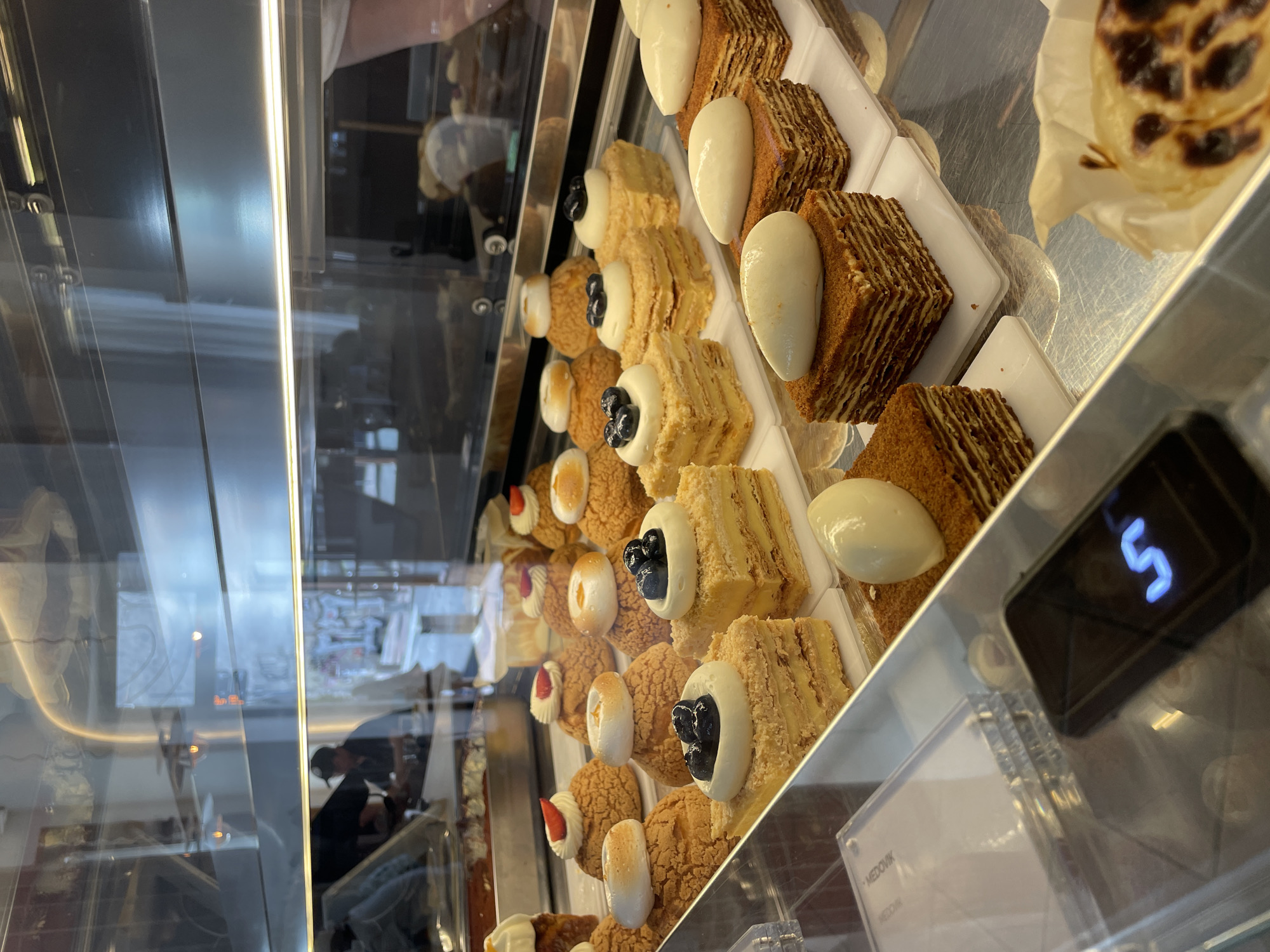 The image shows a display case in a bakery or pastry shop in Belgrade, Serbia. Inside the case, rows of pastries and cakes are arranged on white trays. The pastries include cream puffs topped with fruit, and layered cakes with white cream toppings. The case has a digital display showing the number "5". The interior of the shop is visible through the glass, including a person standing in the background. The pastries and cakes are neatly arranged, suggesting a focus on presentation. The overall impression is that of a clean and well-stocked bakery.