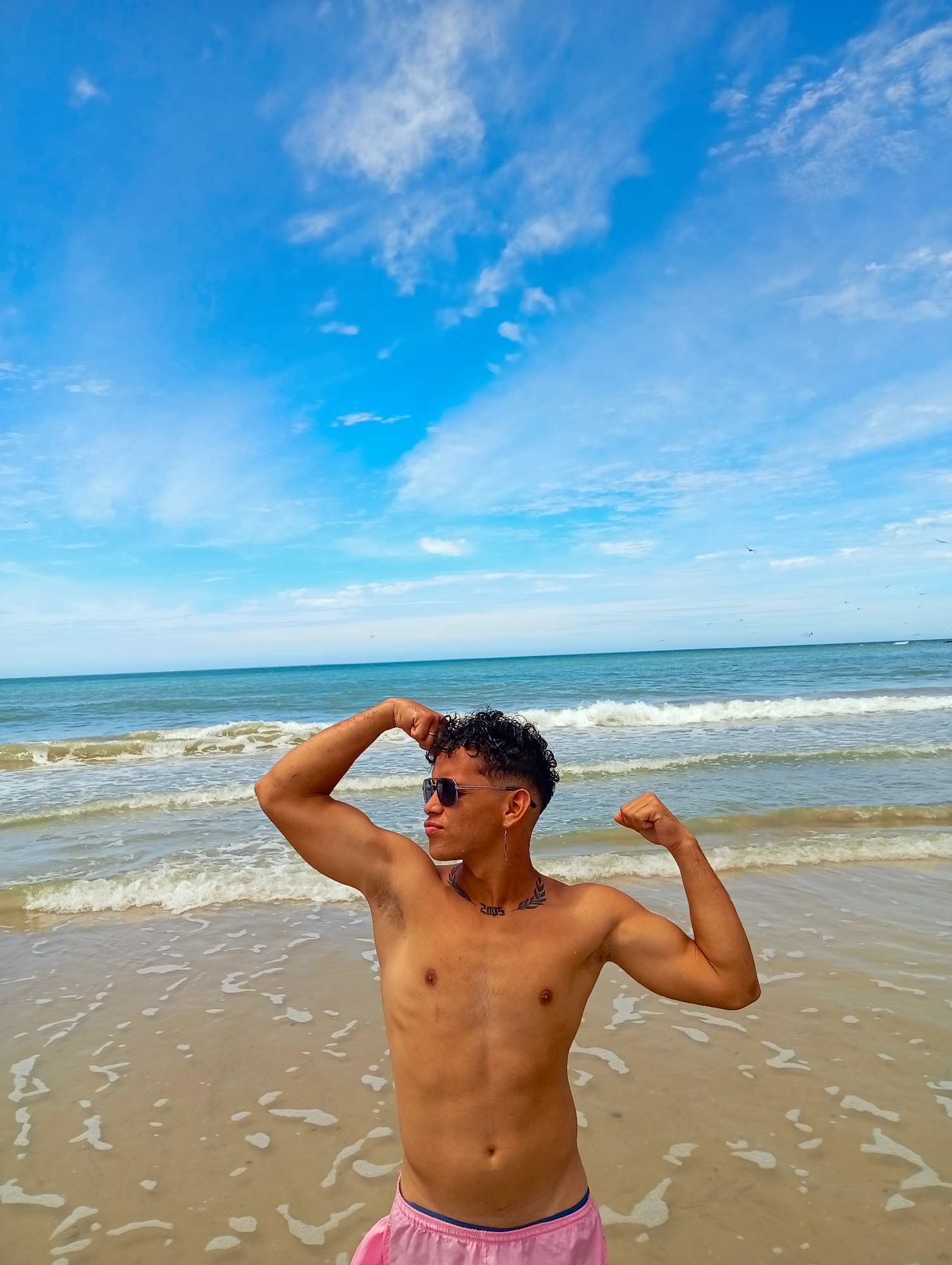 A young man with curly dark hair and sunglasses is standing on a beach. He is flexing his arms, and he has a tattoo on his upper chest. He is wearing pink swim trunks. The beach has light brown sand, and the water is a light blue with white waves. The sky is bright blue with some scattered white clouds. The location is Altagracia, Venezuela.