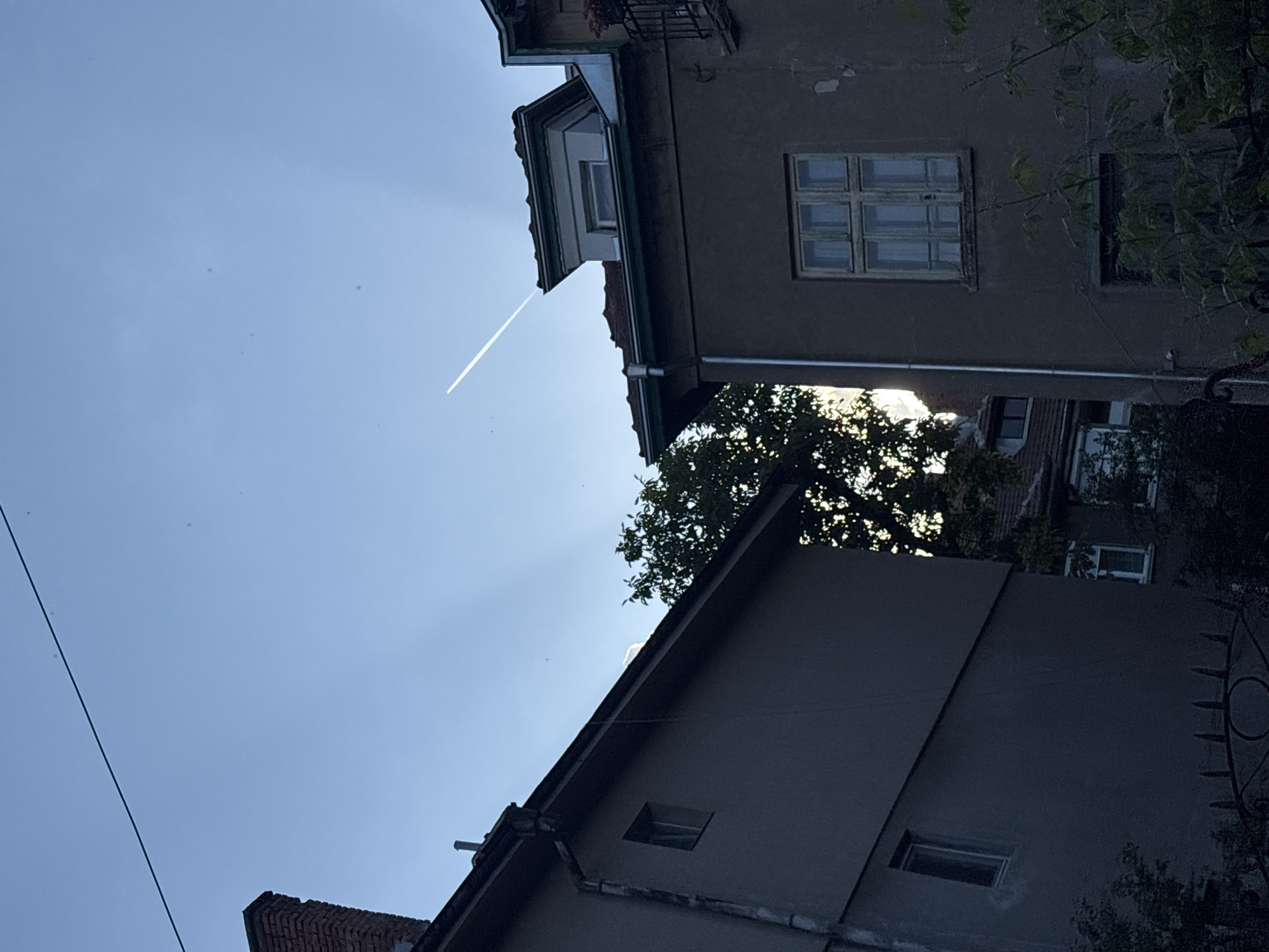 The image depicts a view of a blue sky with a contrail, framed by the rooftops and upper stories of buildings. The buildings, likely residential, are constructed from brick and concrete. Visible details include window frames, gutters, and some foliage. The location is likely Sofia, Bulgaria, based on the urban architecture. An electrical wire is visible in the lower left of the frame.