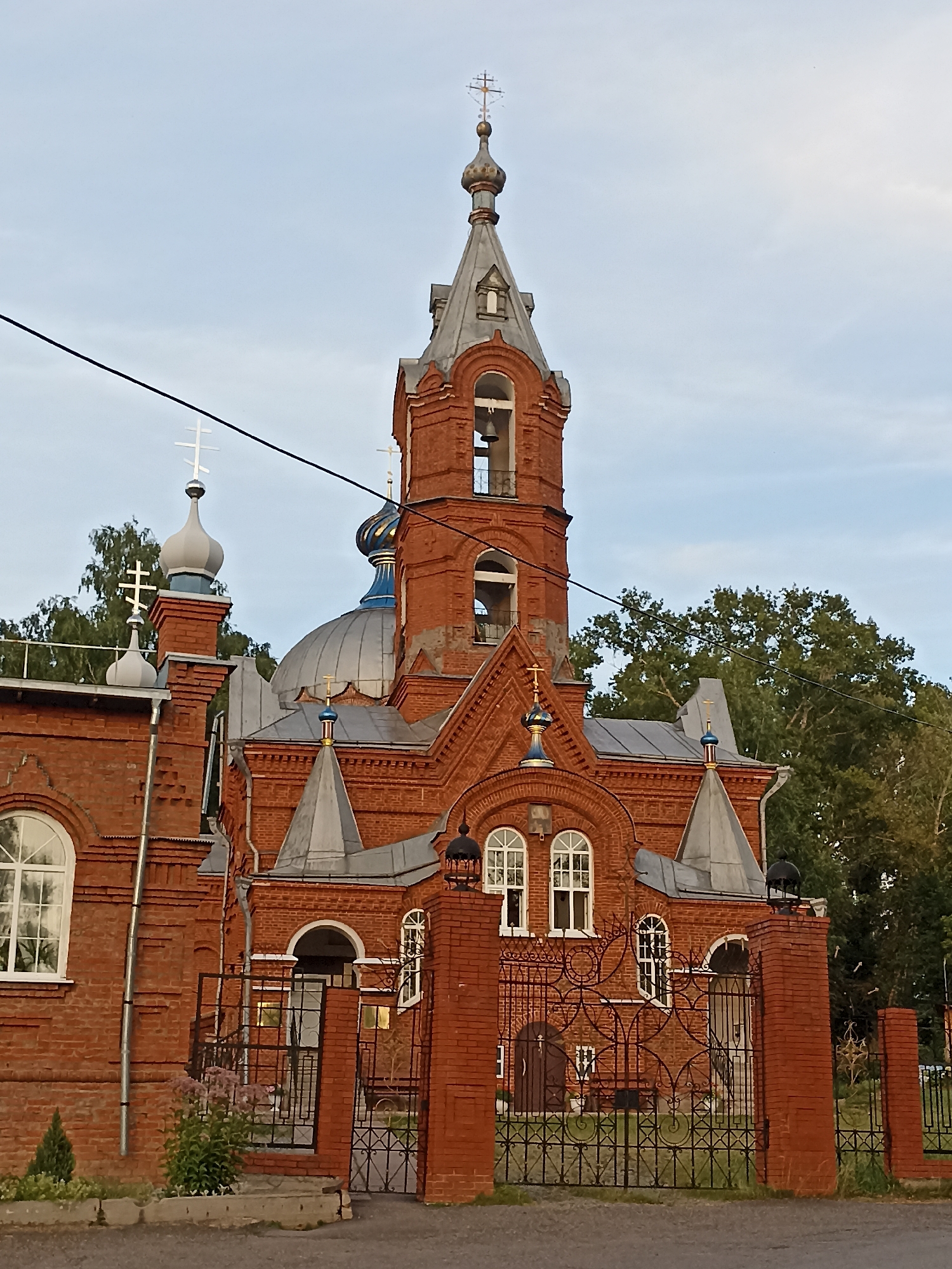 The image shows a red brick Orthodox church in Votkinsk, Russia, on a partly cloudy day. The church has a prominent bell tower with a silver-colored roof and a cross on top. There are also several domes, some of which are blue. The church is surrounded by a metal fence with decorative patterns. Trees are visible in the background, and there is a power line running across the upper part of the image. The entrance to the church is partially obscured by the fence. There are no people visible in the image. The scene appears peaceful and still.