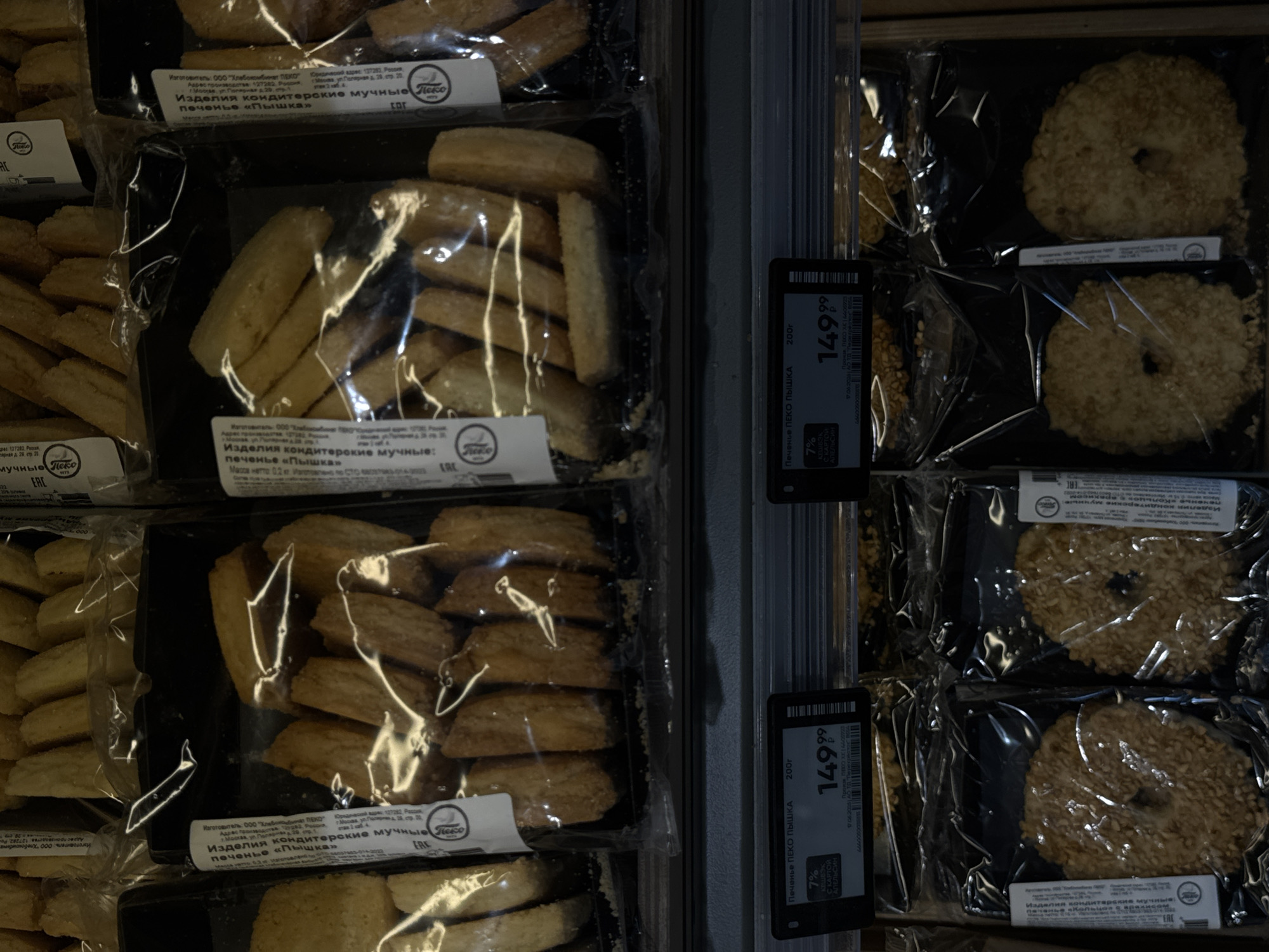 The image shows a retail display of baked goods in Moscow, Russia. In the upper section, various types of cookies are visible in clear plastic packaging. Some cookies are long and rectangular, while others are ring-shaped and coated in sesame seeds. In the lower section, prices are displayed on black price tags. The price tag on the left indicates "Печенье ПЕКО ПЫШКА" (Peko Pyshka Cookies) for 200g, priced at 149.99 rubles. It also highlights a 7% discount for "С КАРТОЙ АПЕЛЬСИН" (with Apelsin card). The environment appears to be a supermarket or bakery aisle, with goods stacked on shelves. The lighting is artificial, typical of an indoor retail setting. No people are visible in the frame.