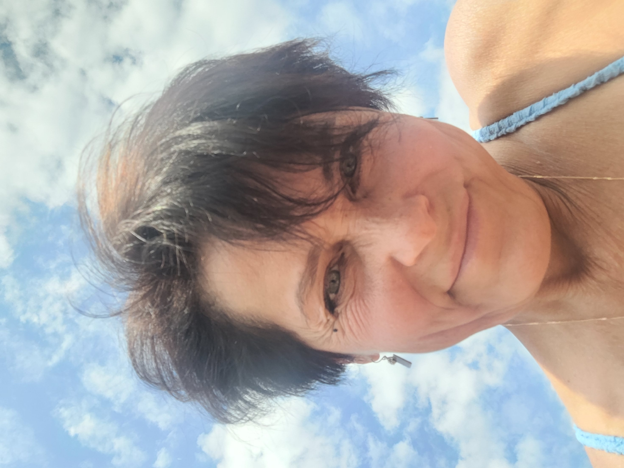 A woman with short, dark hair and light-colored eyes smiles at the camera. She is wearing a light blue top with a textured strap. Her left earlobe is adorned with a small, rectangular earring. The background is a bright blue sky filled with scattered white clouds. The image appears to have been taken outdoors, possibly on a sunny day. It is not possible to determine the exact location within Zelenogorsk, Russia based on the provided image.