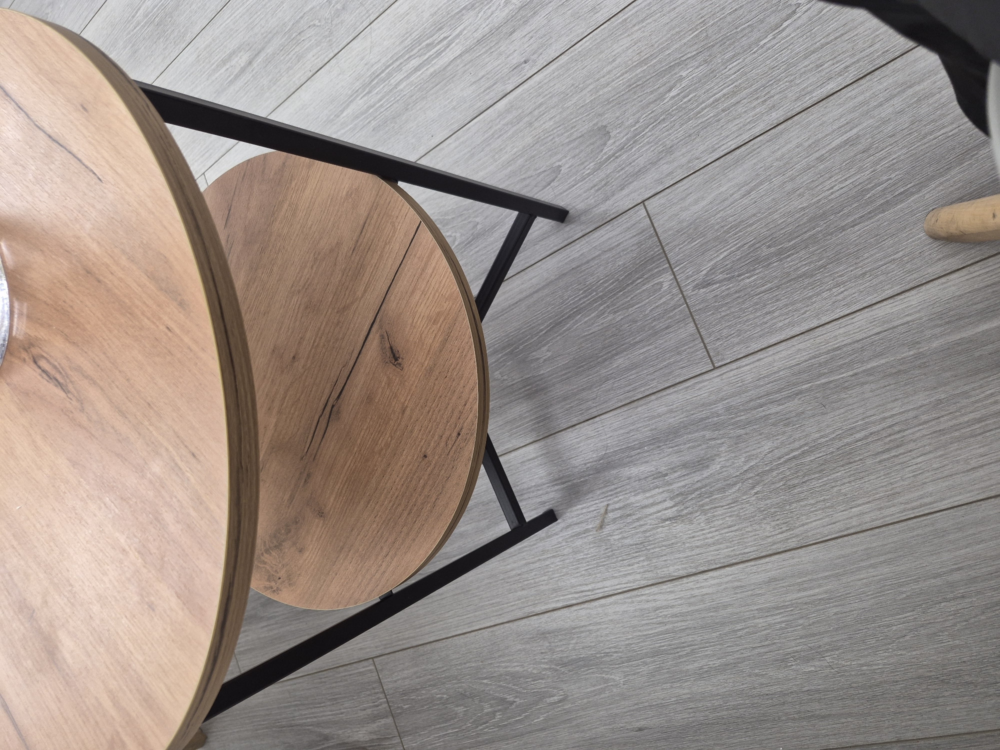 This close-up, high-angle shot captures a modern interior scene, set in what is contextualized as Yerevan, Armenia. The primary subjects are two circular table tops, partially visible, made of light-colored wood with distinct grain patterns and some darker, natural-looking imperfections or knots. These tables appear to be part of a nesting set or side tables, with the larger one closer to the viewer and the smaller one positioned slightly behind and to its right. They are supported by a sleek black metal frame with angular lines, visible beneath and between the table tops.
The floor, which covers a significant portion of the image, is composed of grey wooden planks, likely laminate or engineered wood. These planks are arranged diagonally across the frame, exhibiting a fine wood texture with parallel lines. In the bottom right corner, a small, partial view of a light-colored, possibly wooden, leg from another piece of furniture (such as a chair) is visible. A dark, indistinct object or fabric can also be seen along the top right edge of the frame.
There is no specific activity taking place; the image captures a static arrangement of furniture. No visible text is present in the image, nor are there cues to indicate the time of day or specific weather conditions. The photograph emphasizes the textures of the wood and flooring, along with the minimalist design of the tables.