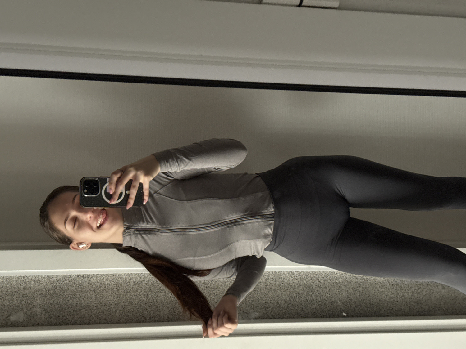 An indoor mirror selfie captures a young woman from the chest down, rotated 90 degrees in the original image but depicted as upright here. She is standing in front of a large mirror, holding a dark-colored smartphone to take her picture. The woman has long, dark brown hair styled in a ponytail, which she is holding with her left hand near her hip. She is wearing a light grey, long-sleeved athletic top with a full zipper down the front and dark grey or black form-fitting athletic leggings. White earbuds are visible in her ears, and a ring is on her right hand, which is holding the phone. Her facial expression is a playful smile with her tongue slightly extended. The smartphone case features a distinct white circular design, possibly a peace sign. The background reflects a plain light-colored wall and what appears to be a grey, textured floor, possibly carpet or a rug, with a white baseboard or frame visible at the bottom. The setting is likely a private room or hallway in Sofia, Bulgaria.