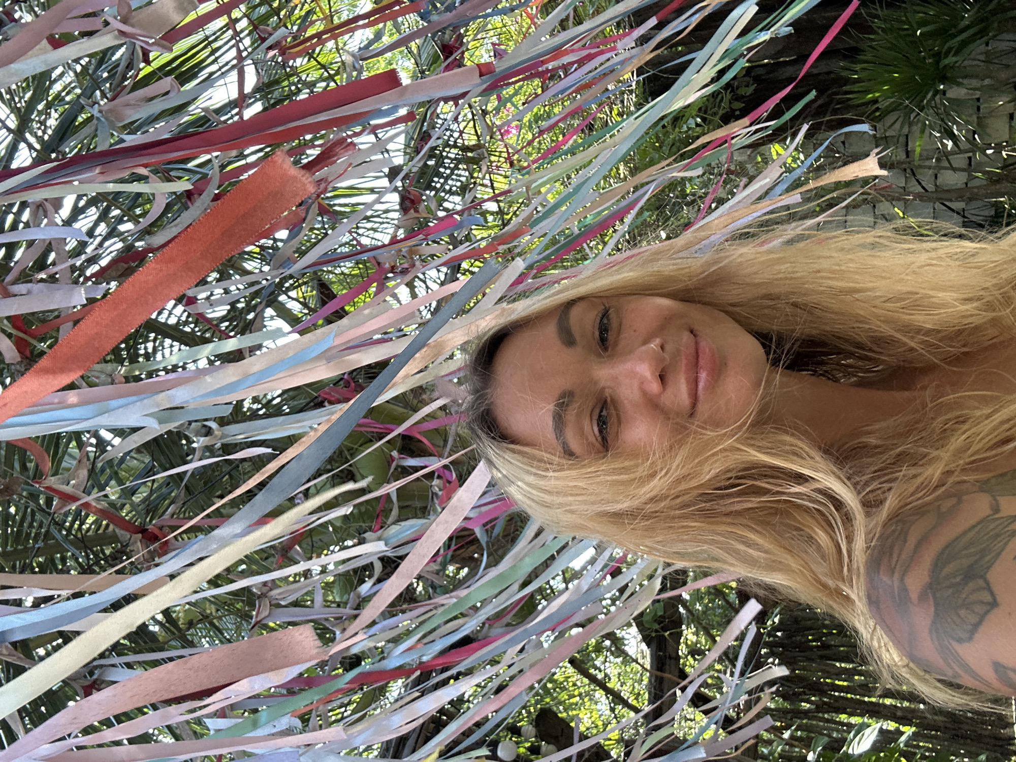The image shows a portrait of a young blonde woman with a tattoo on her left shoulder, smiling at the camera. She is positioned in the foreground, with a background of palm fronds and colorful ribbons hanging from trees. The scene appears to be outdoors, possibly in a tropical setting. The woman's hair is windswept, and she is wearing a light-colored top that is mostly out of frame. The ribbons are of various colors including pink, blue, white, and red, and they are blowing in the breeze, creating a festive atmosphere. The overall impression is one of a relaxed and joyful moment captured in a vibrant, natural environment. It is impossible to definitively determine the specific location in Mũi Né, Vietnam, based solely on the visual information provided in the image.