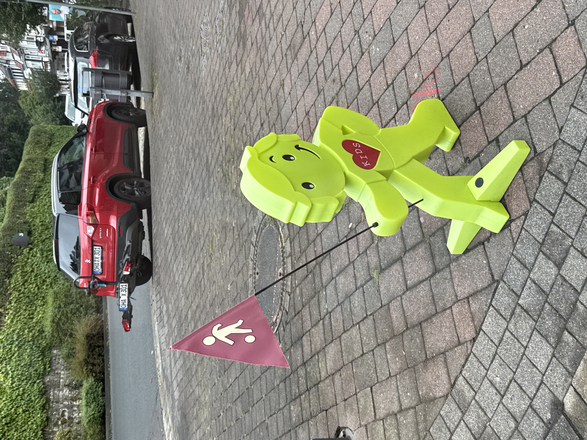 An outdoor, eye-level shot captures a bright lime green plastic figure resembling a stylized child, positioned on a cobblestone sidewalk. The figure, which has a simple smiley face, is holding a dark thin pole with a small pink triangular flag attached. The flag features a white silhouette of a person kicking a soccer ball. The figure's chest is emblazoned with a red heart containing the white word "KIDS."
In the background, a red Suzuki Vitara SUV is parked on the street. The SUV has a bicycle rack attached to its rear, and its license plate reads "OHA X 301." To the right of the SUV, a dark colored car is parked behind it. The street is lined with houses and trees. The area appears to be in Bad Sachsa, Germany, as indicated by the license plate format and the architectural style of the buildings. The overall scene suggests a residential or public area where children might be present.