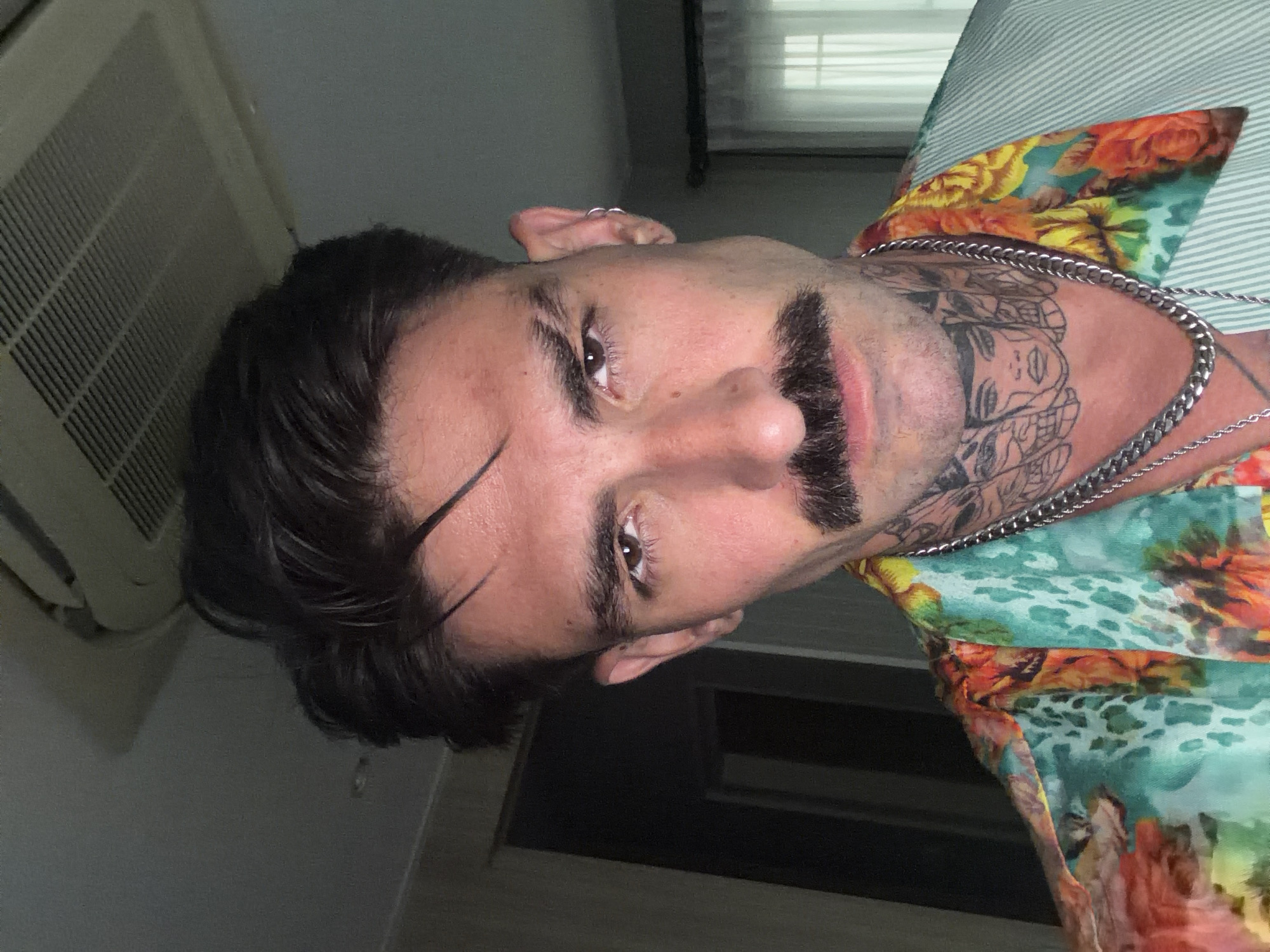 A medium close-up shot depicts a light-skinned individual with dark hair, a single prominent eyebrow slit, a thick mustache, and visible neck tattoos, positioned slightly askew relative to the camera's perspective. The person is wearing a colorful floral-patterned shirt and multiple silver chain necklaces. The environment includes an air conditioning unit in the upper left corner and a glimpse of a window with blinds. The location is indeterminable to be Chalong, Thailand.