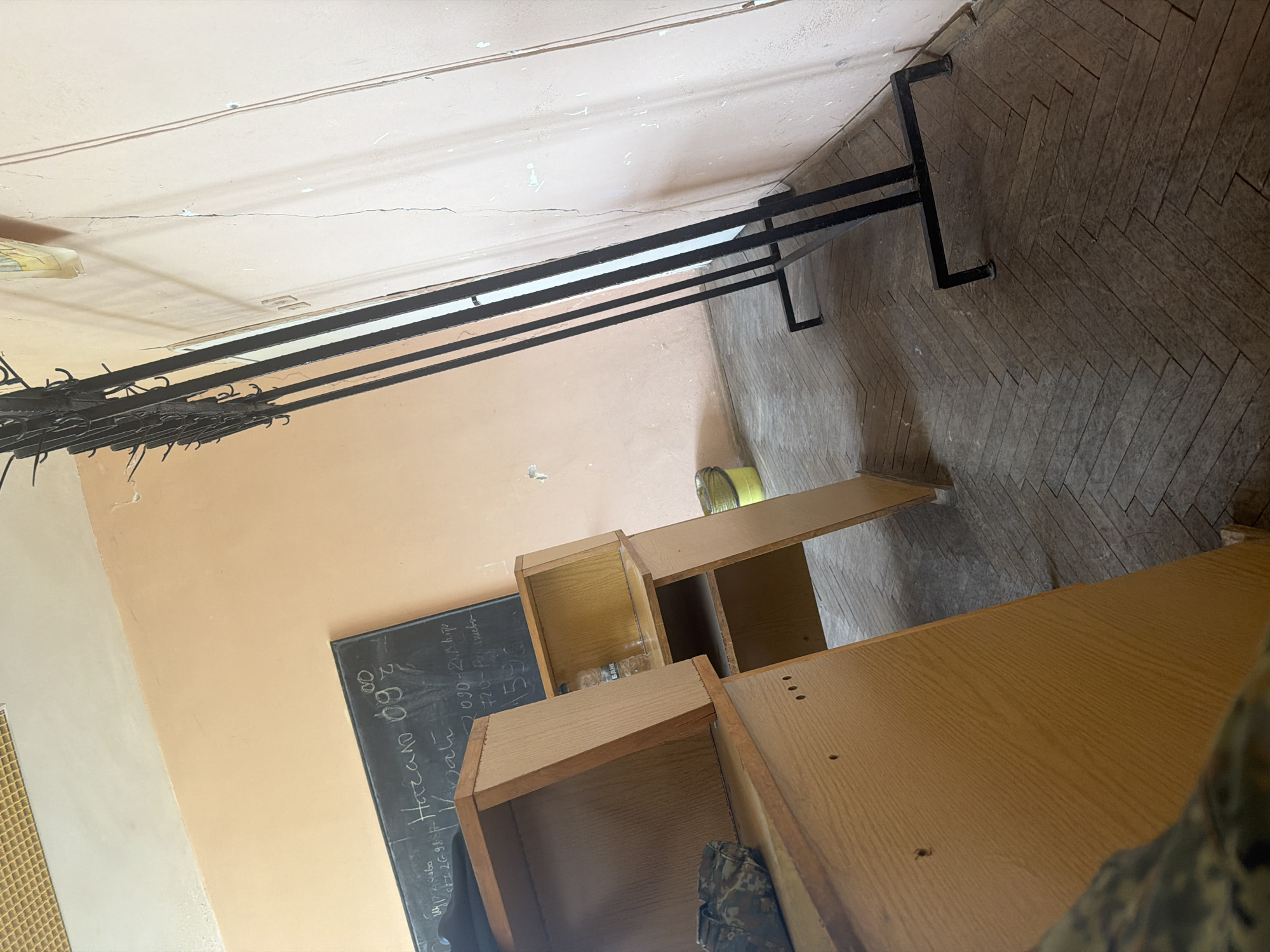 The image shows an interior view, potentially a classroom or office space, in Dolna Mitropolia, Bulgaria. The walls are light orange, and the floor is covered in a herringbone pattern of light brown tiles. Visible in the room are several wooden cabinets or shelving units, a black chalkboard with handwriting in white chalk, and a black metal clothing rack attached to the wall near the ceiling. There are no people explicitly visible, but there may be an individual partially visible in the lower right corner of the image.
